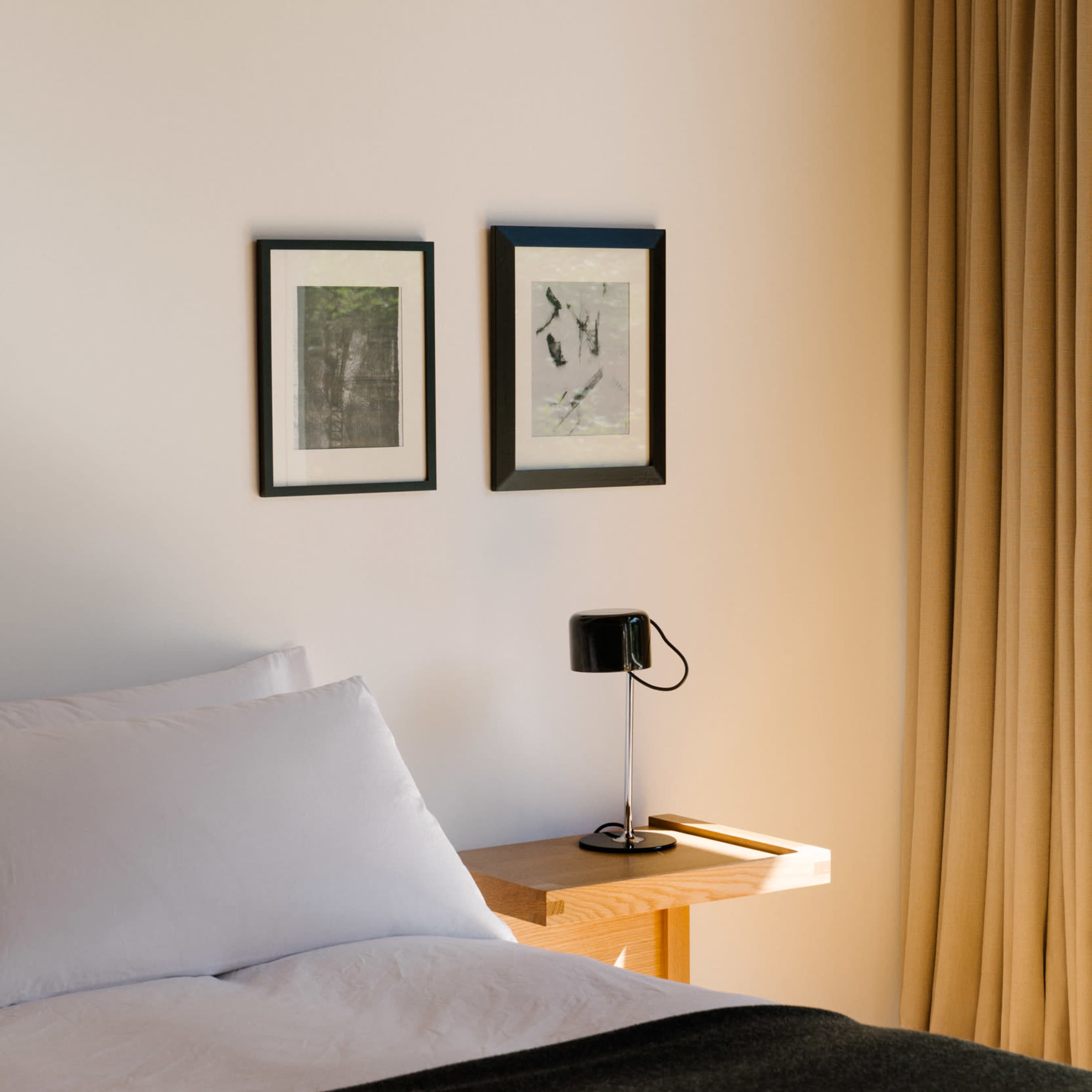 A neatly made bed with white linens, a black bedside lamp, and two framed artworks hanging on the wall beside a window with beige curtains.