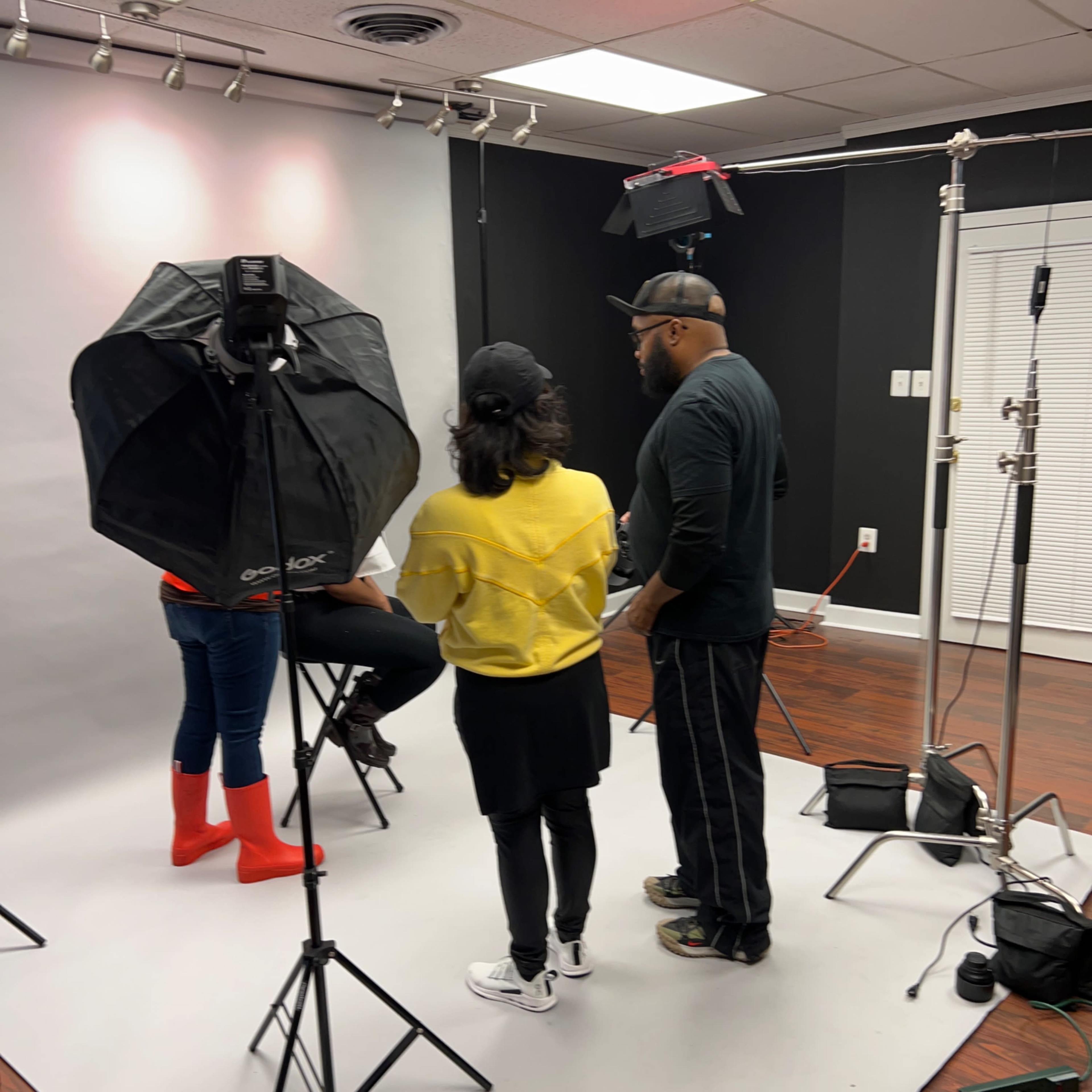 Three people are preparing for a photoshoot in a studio with a gray backdrop and various lighting equipment.