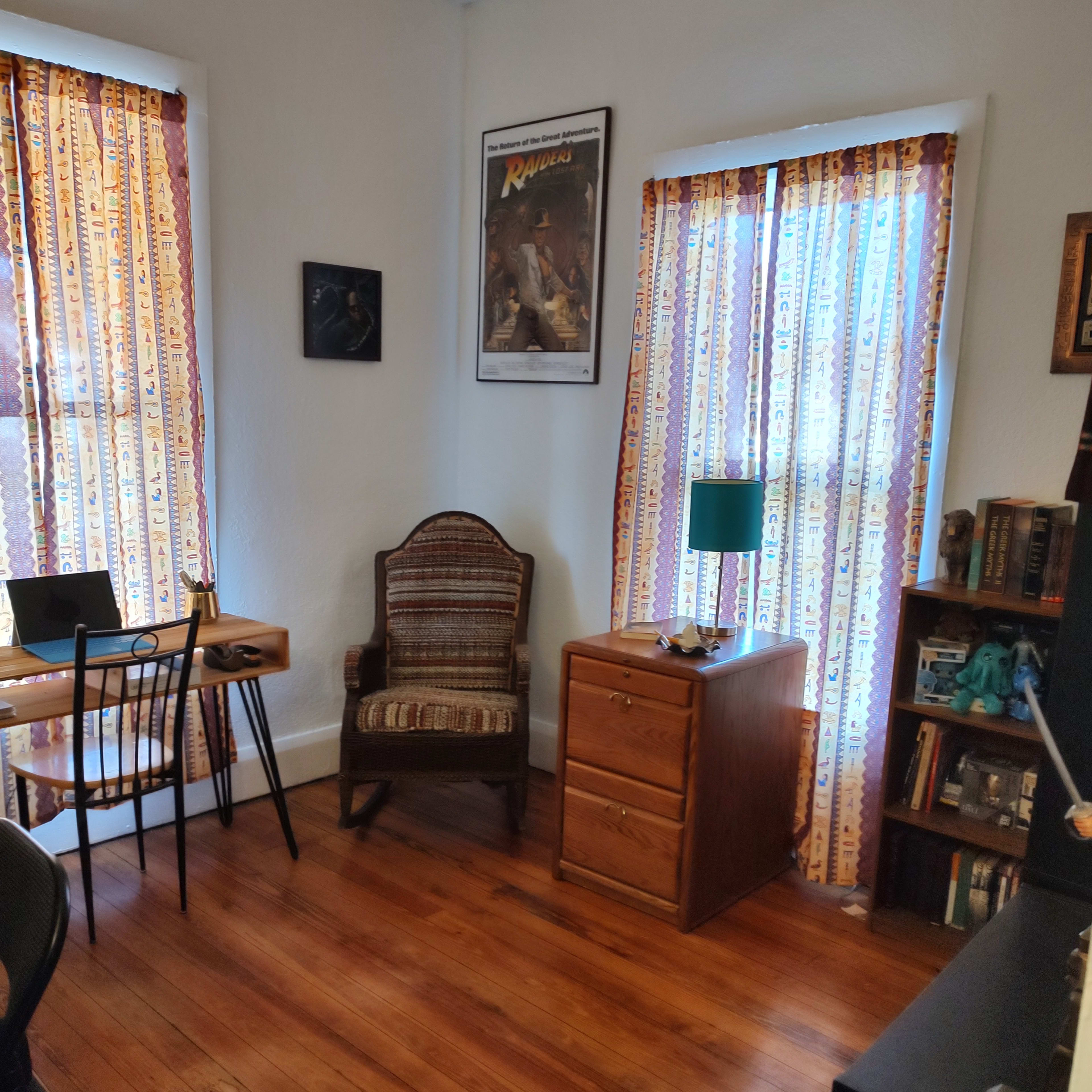 A cozy room with two windows draped in patterned curtains, a wooden chair, a small desk, a lamp on a nightstand, and a bookshelf against the wall.