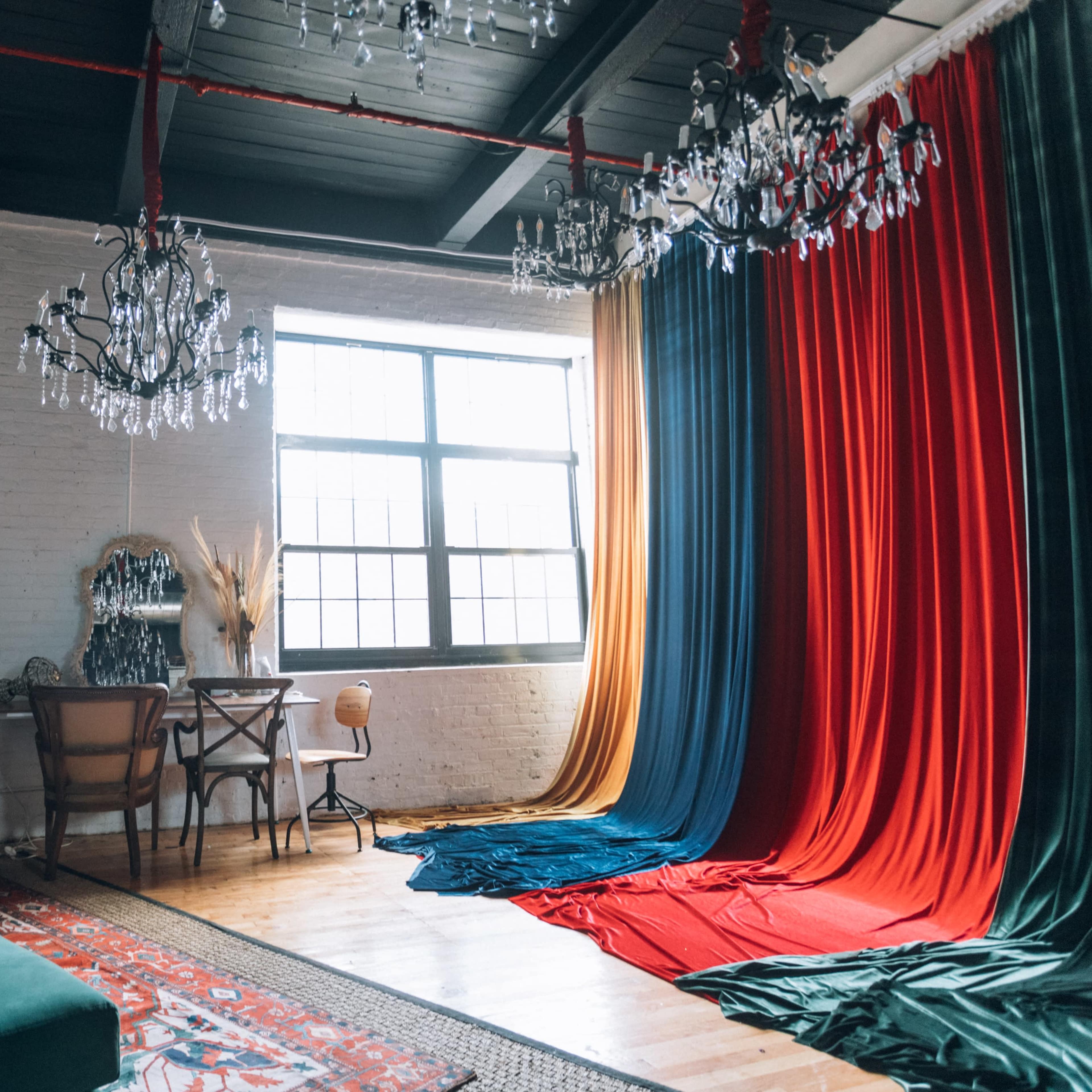 A brightly lit room features a large window, two chandeliers, and colorful fabric drapes in red, blue, and orange alongside a vintage armchair and a small dining table.