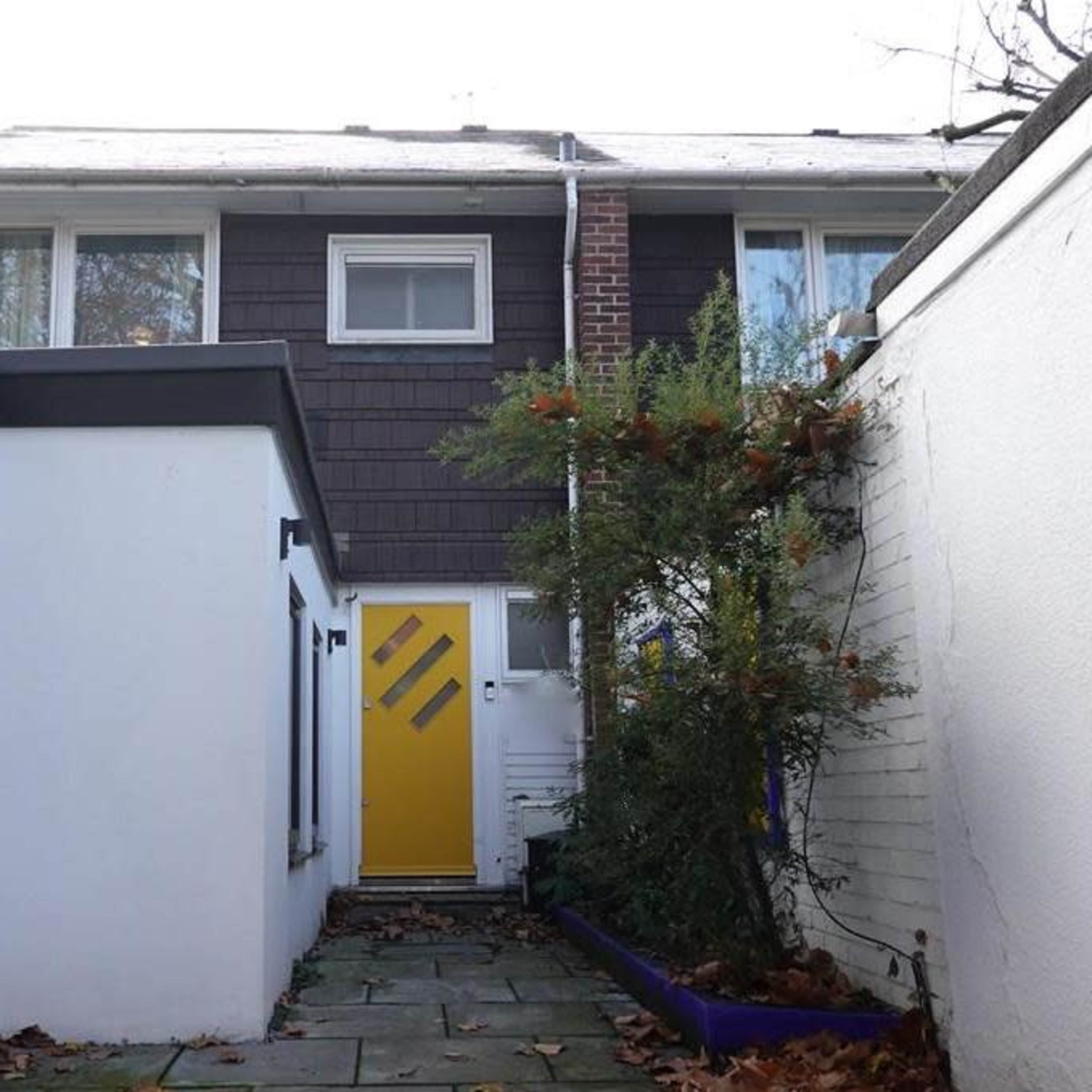 A narrow pathway leading to a yellow door of a house, flanked by white walls and a small garden area.