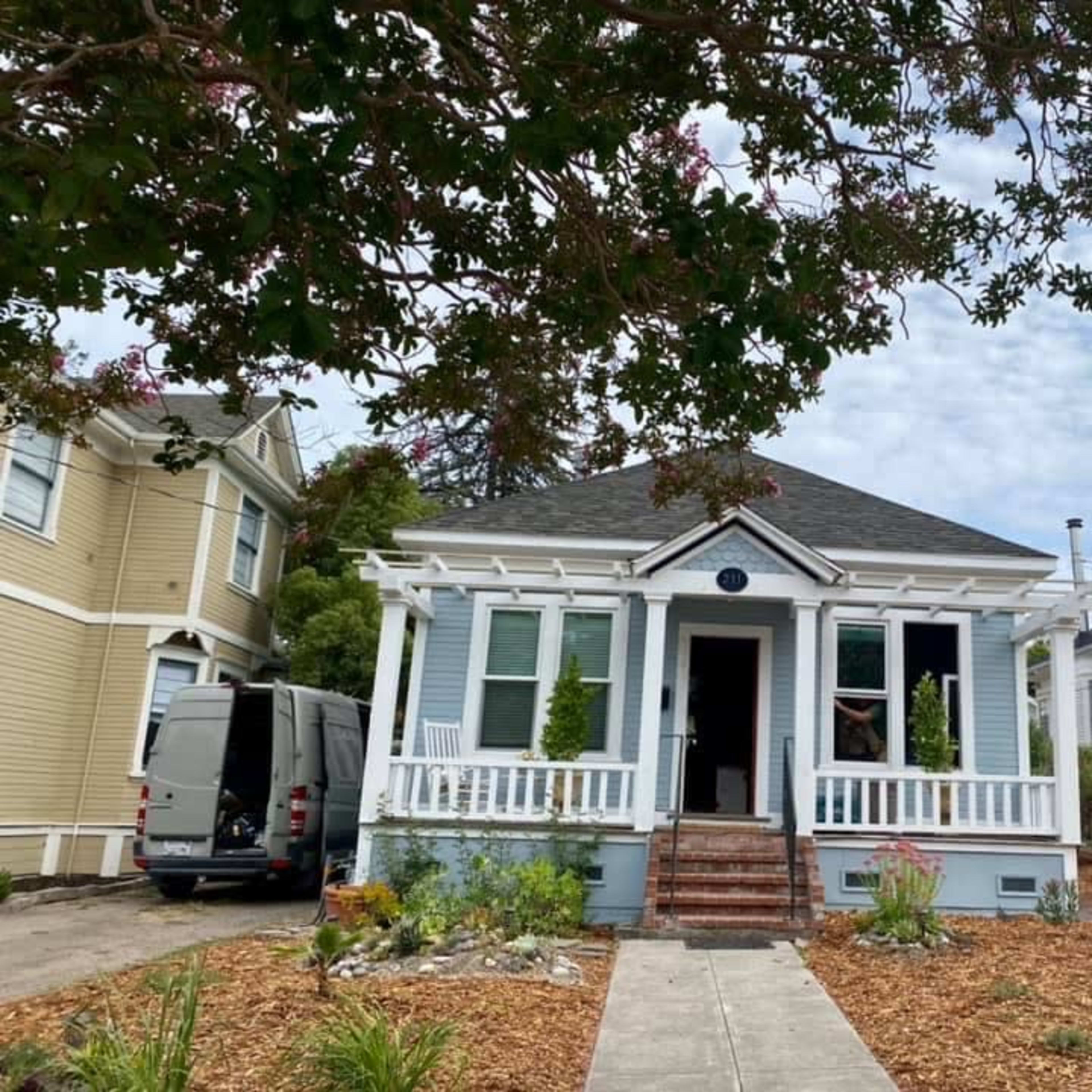 A single-story house with a porch is flanked by two other buildings and surrounded by a landscaped yard.
