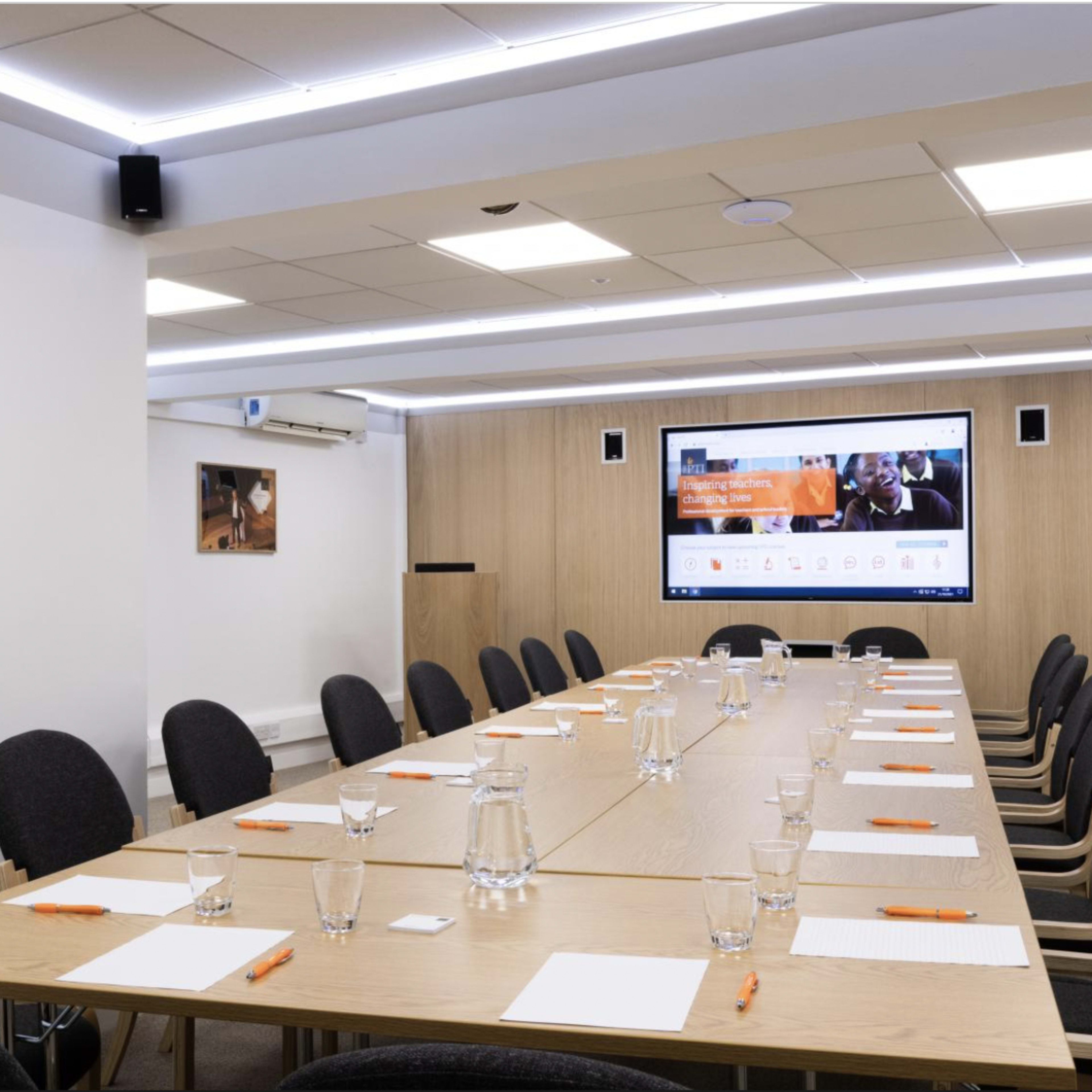 A modern conference room features a long wooden table set with notebooks and pens, facing a large screen displaying digital content.