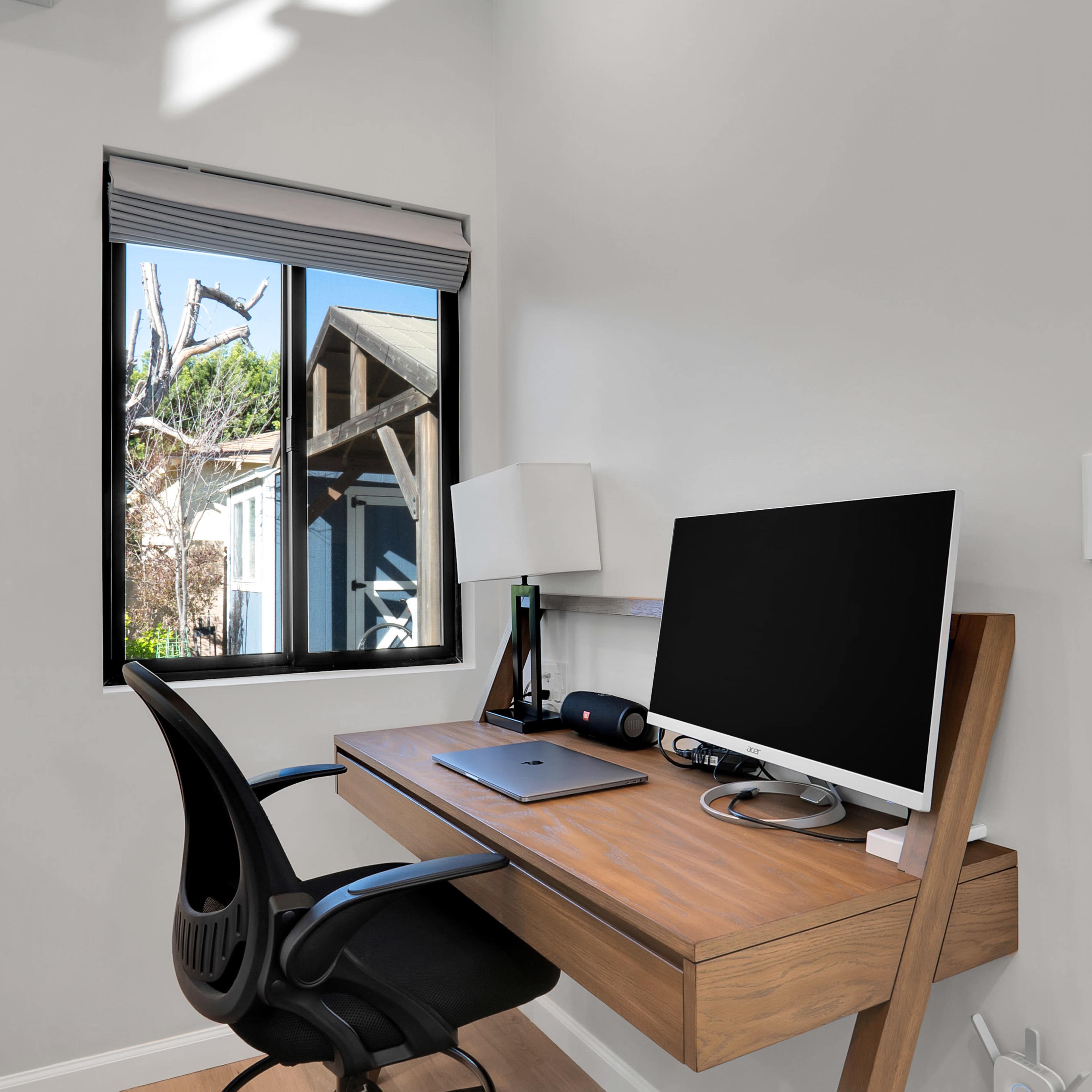 A modern home office features a wooden desk with a computer monitor, an adjustable chair, and a lamp, set beside a window that looks out onto a garden.