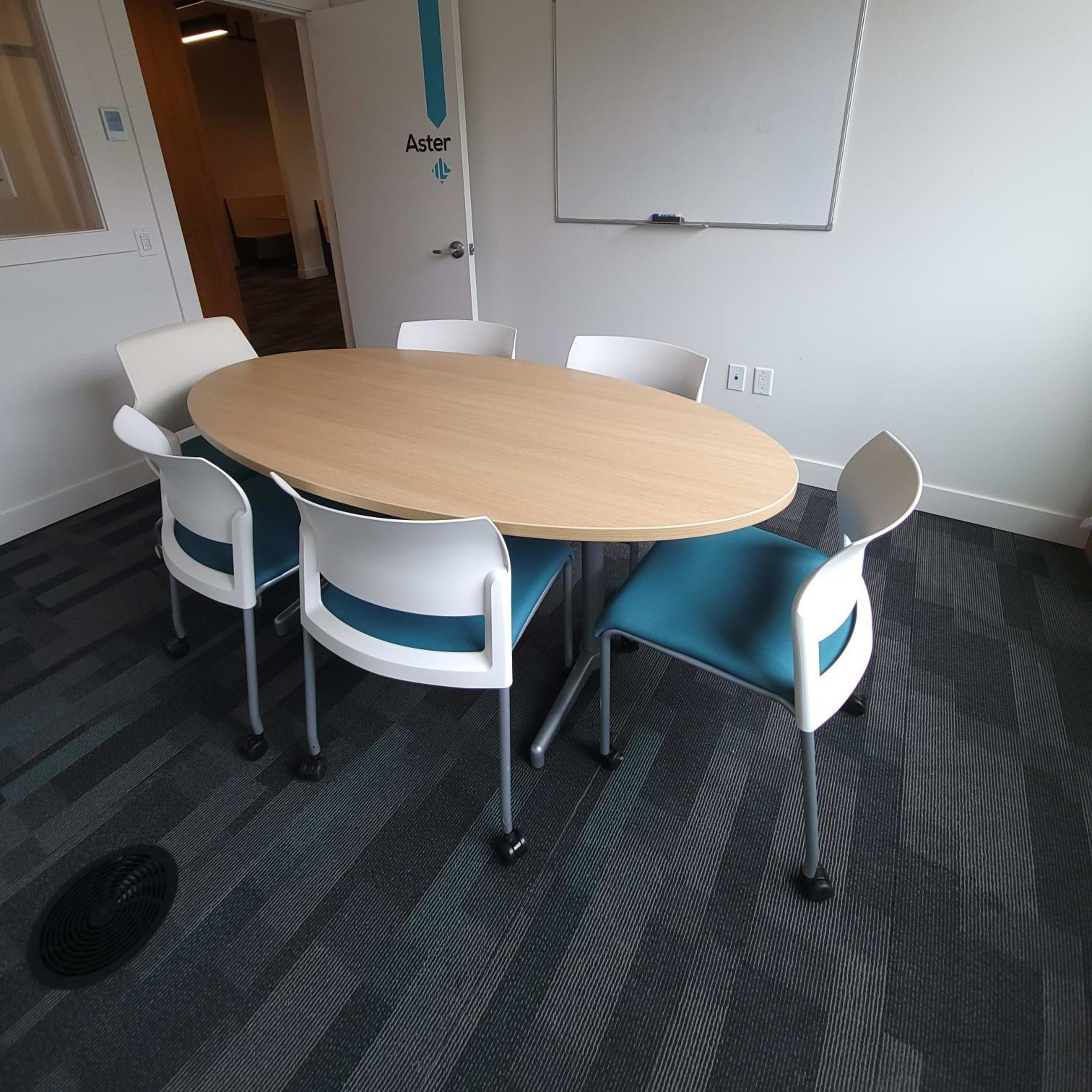 A round wooden table surrounded by five white and blue chairs in a conference room with a whiteboard and a door labeled "Aster."