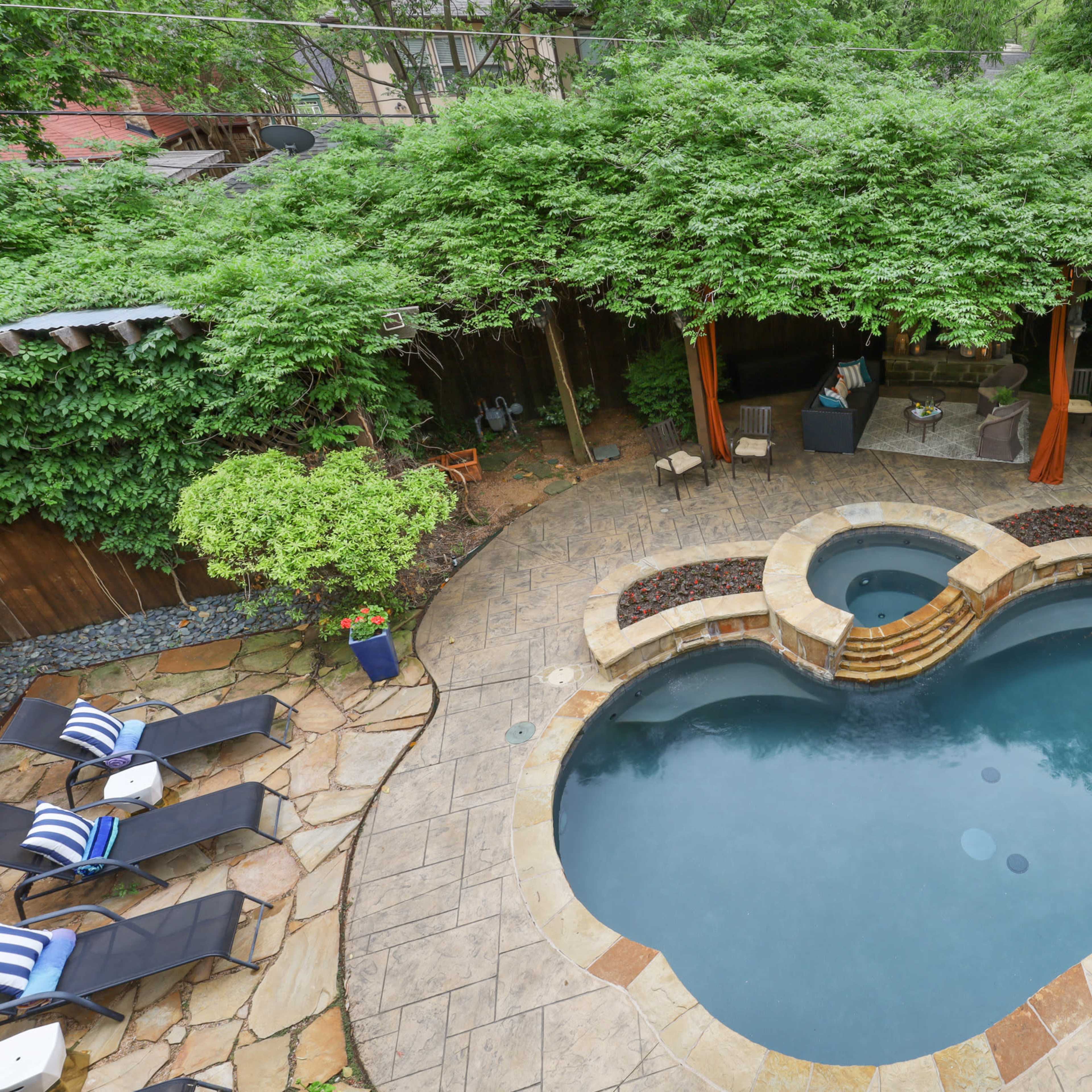 A kidney-shaped swimming pool surrounded by stone patio, lush greenery, and lounge chairs, with a shaded seating area nearby.