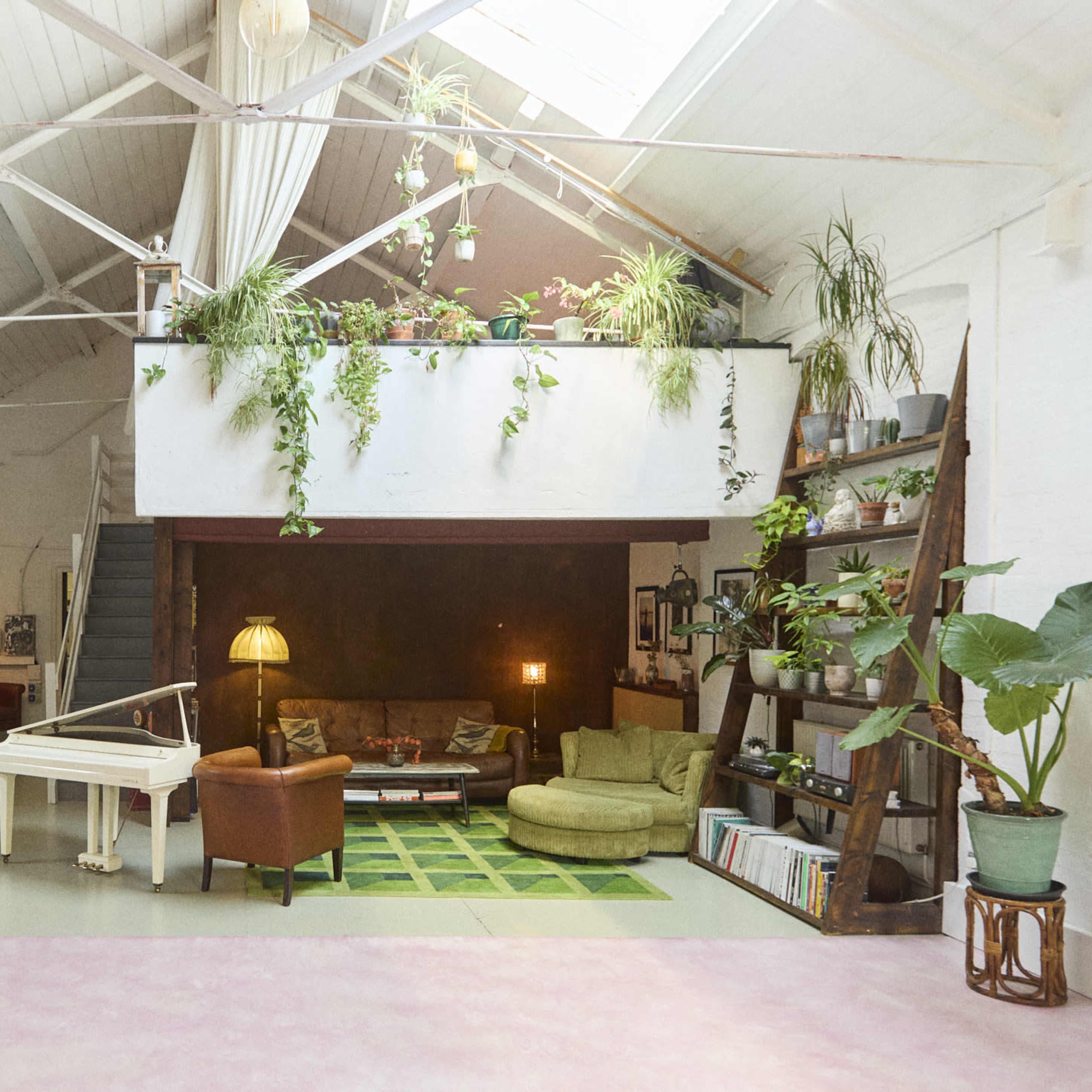 A spacious, brightly lit loft with green plants, a white piano, a green sofa, and shelves filled with books and decorative items.