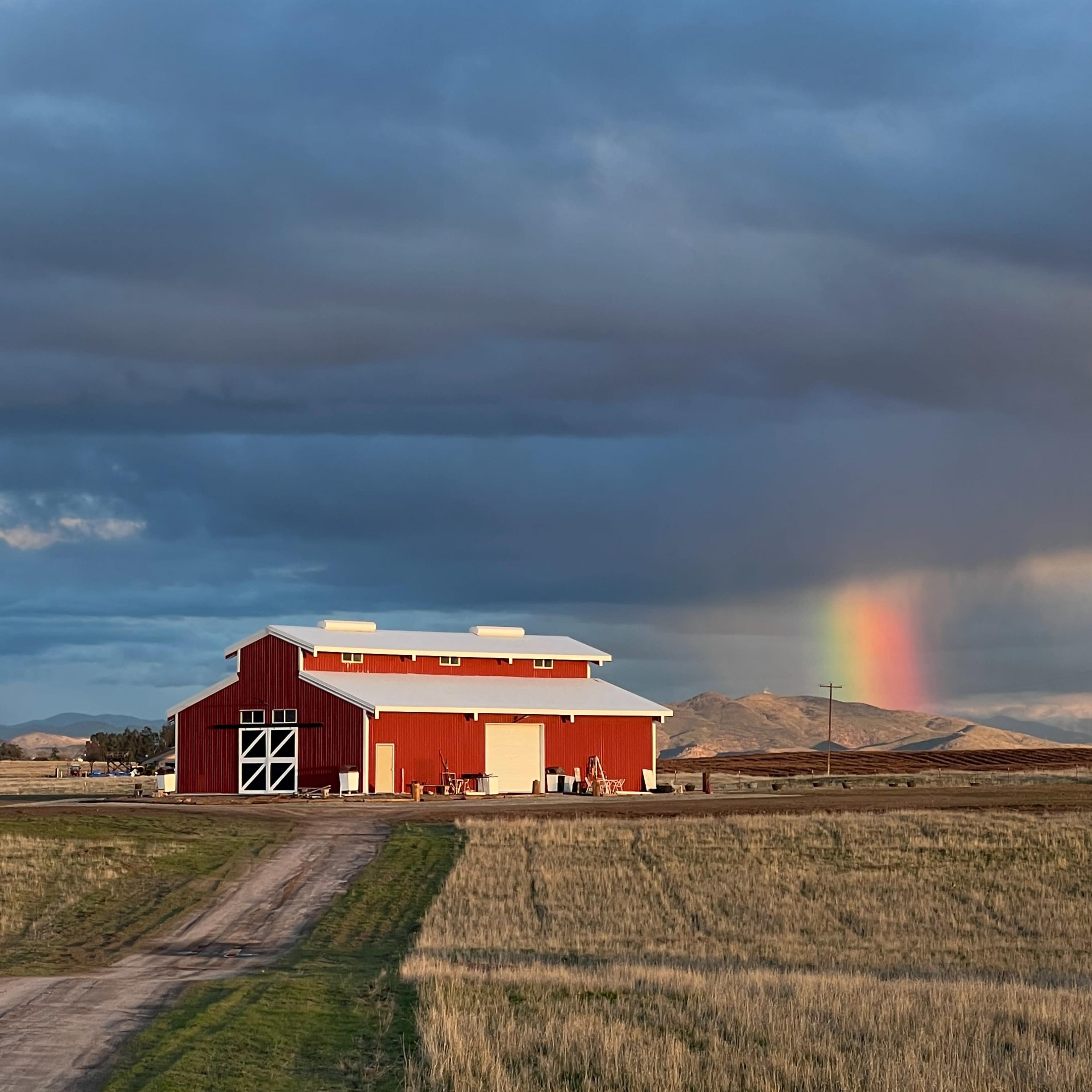 A red barn stands in a field under a cloudy sky, with a rainbow appearing in the distance.