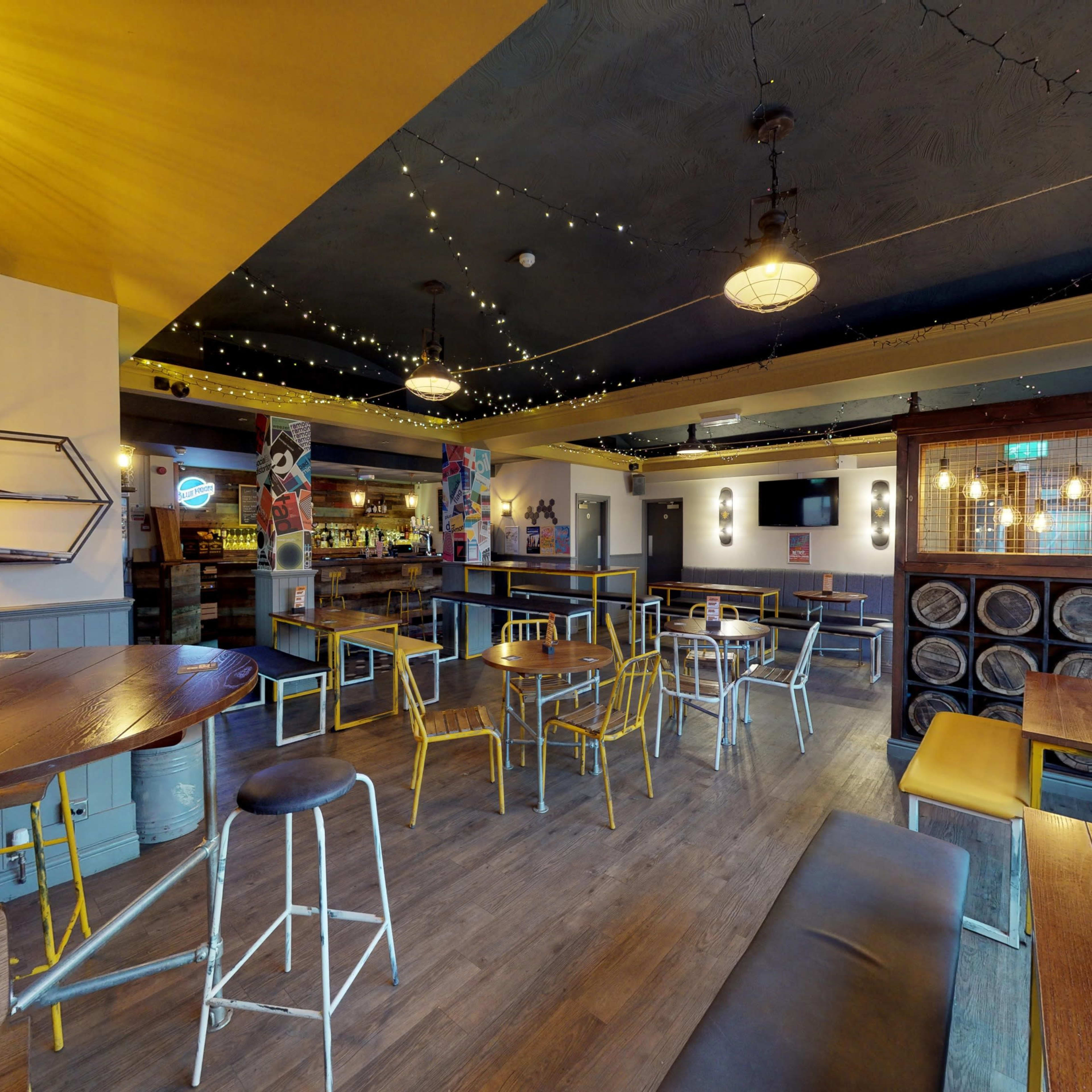 An empty restaurant interior with wooden tables, metal chairs, and decorative lighting, featuring a bar area in the background.