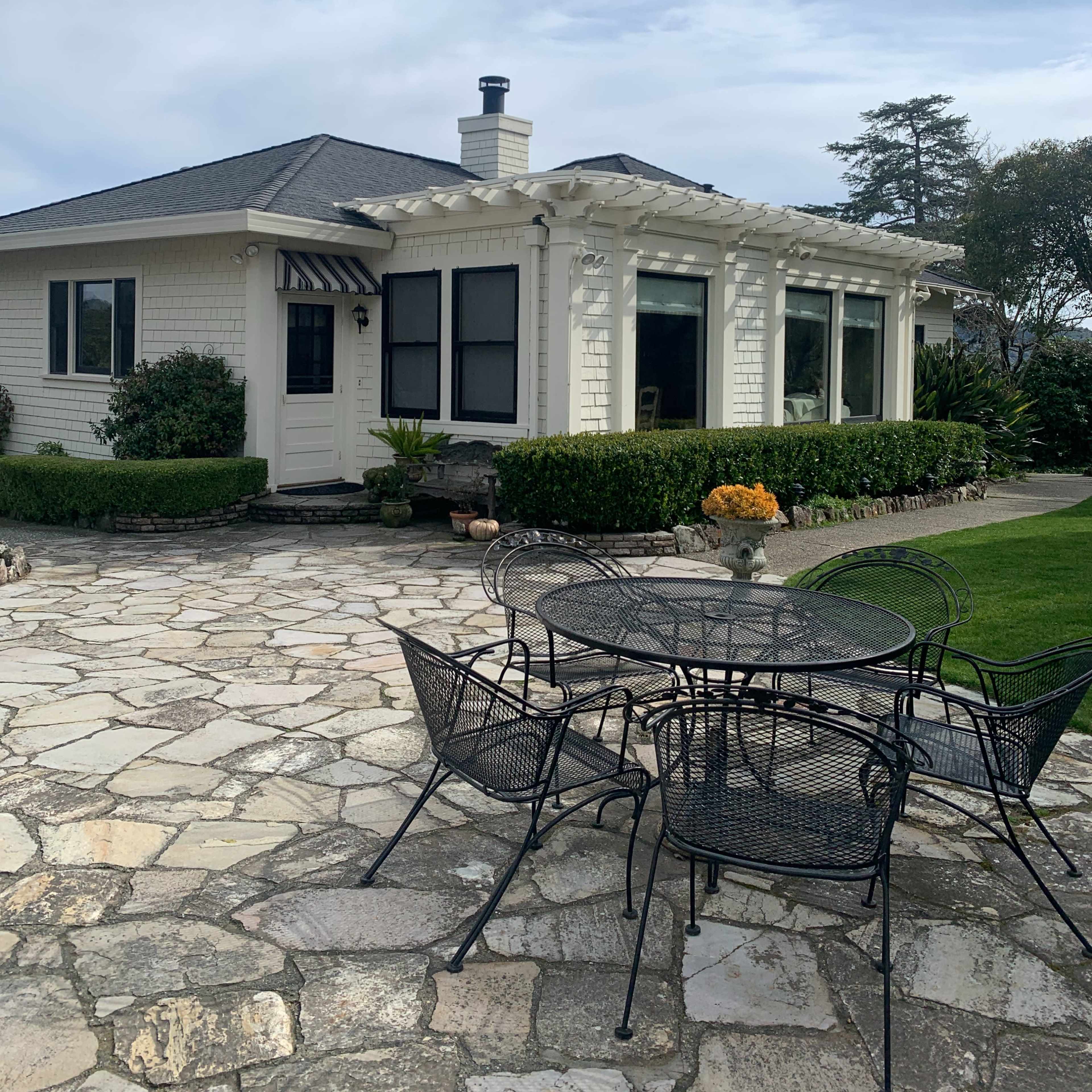 A white house with a porch and large windows is set against a stone patio featuring a black metal table and chairs, surrounded by greenery.