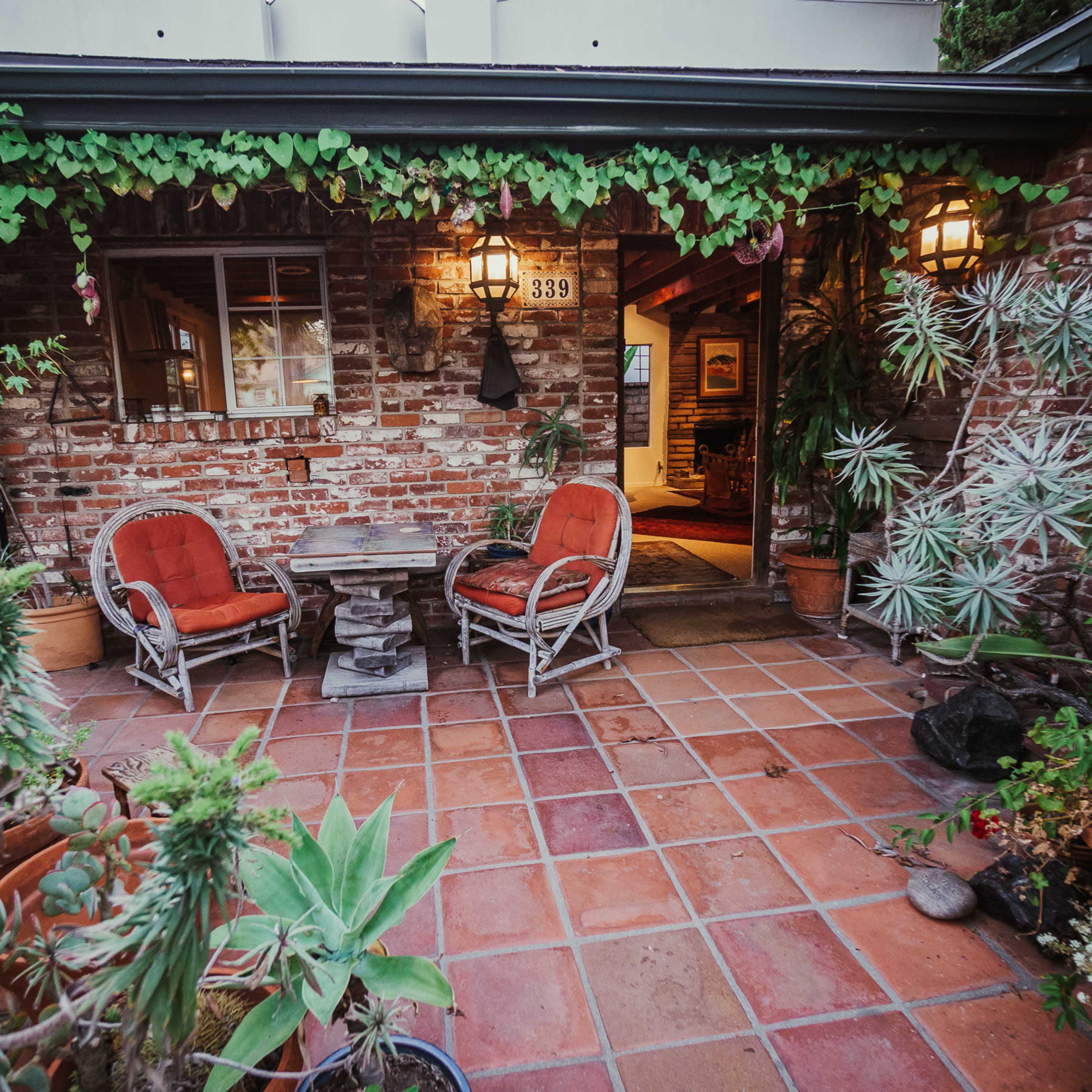 A brick patio area features a small table with two red chairs surrounded by potted plants and vines.
