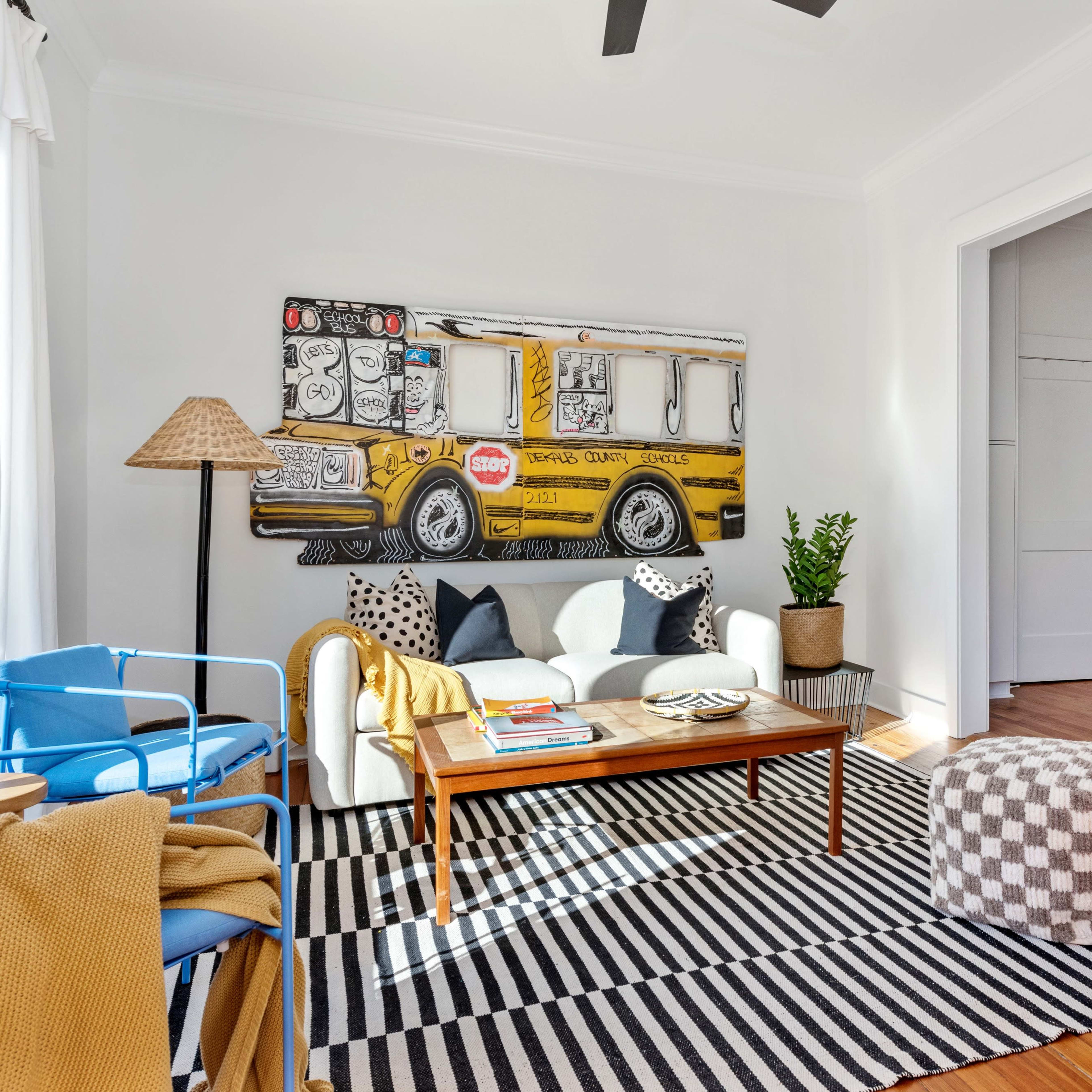 A bright living room features a large bus mural on the wall, a striped rug, a coffee table with books, and seating that includes a blue chair and a sofa adorned with decorative pillows.