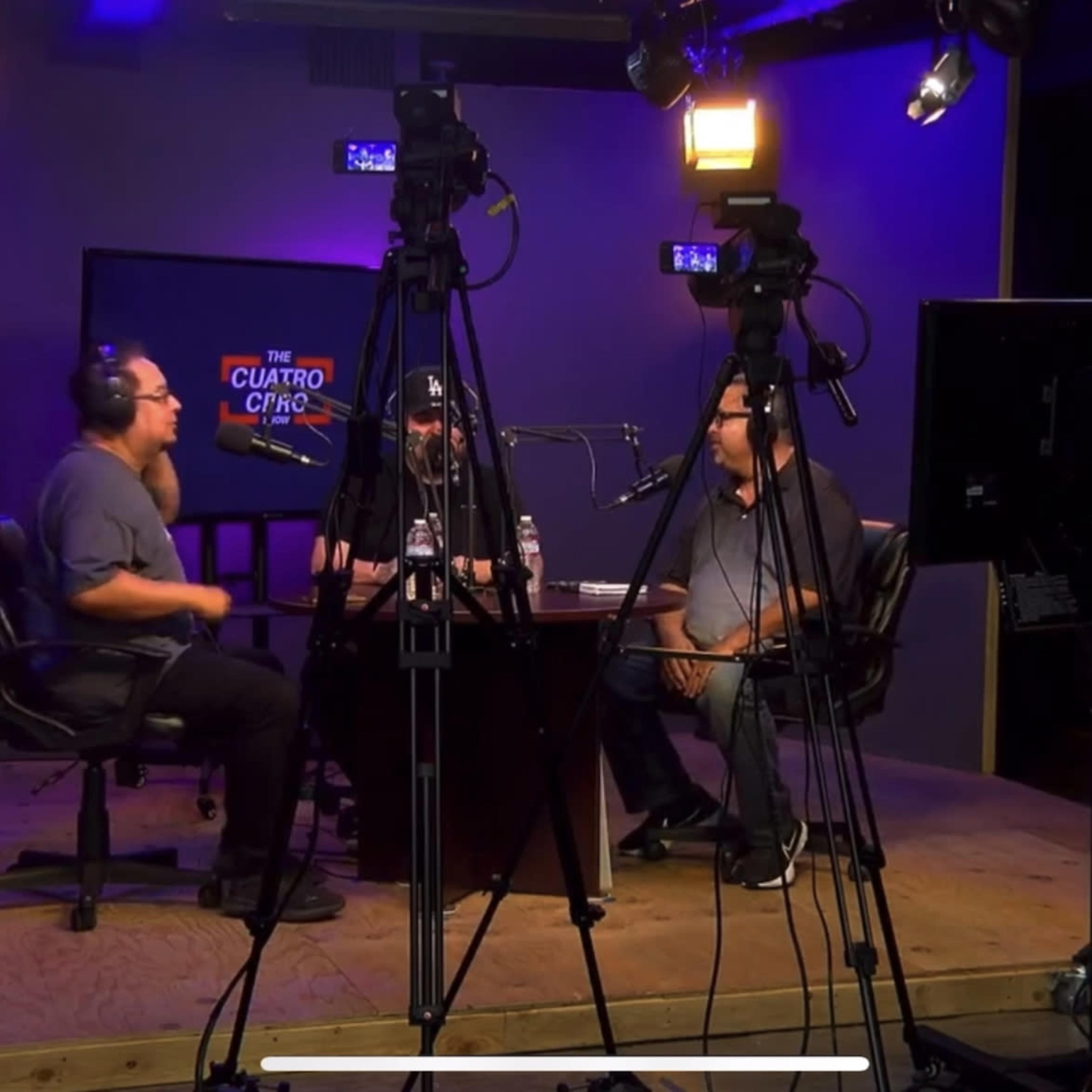 Three individuals are seated around a table in a studio, discussing while surrounded by multiple cameras and studio lights.