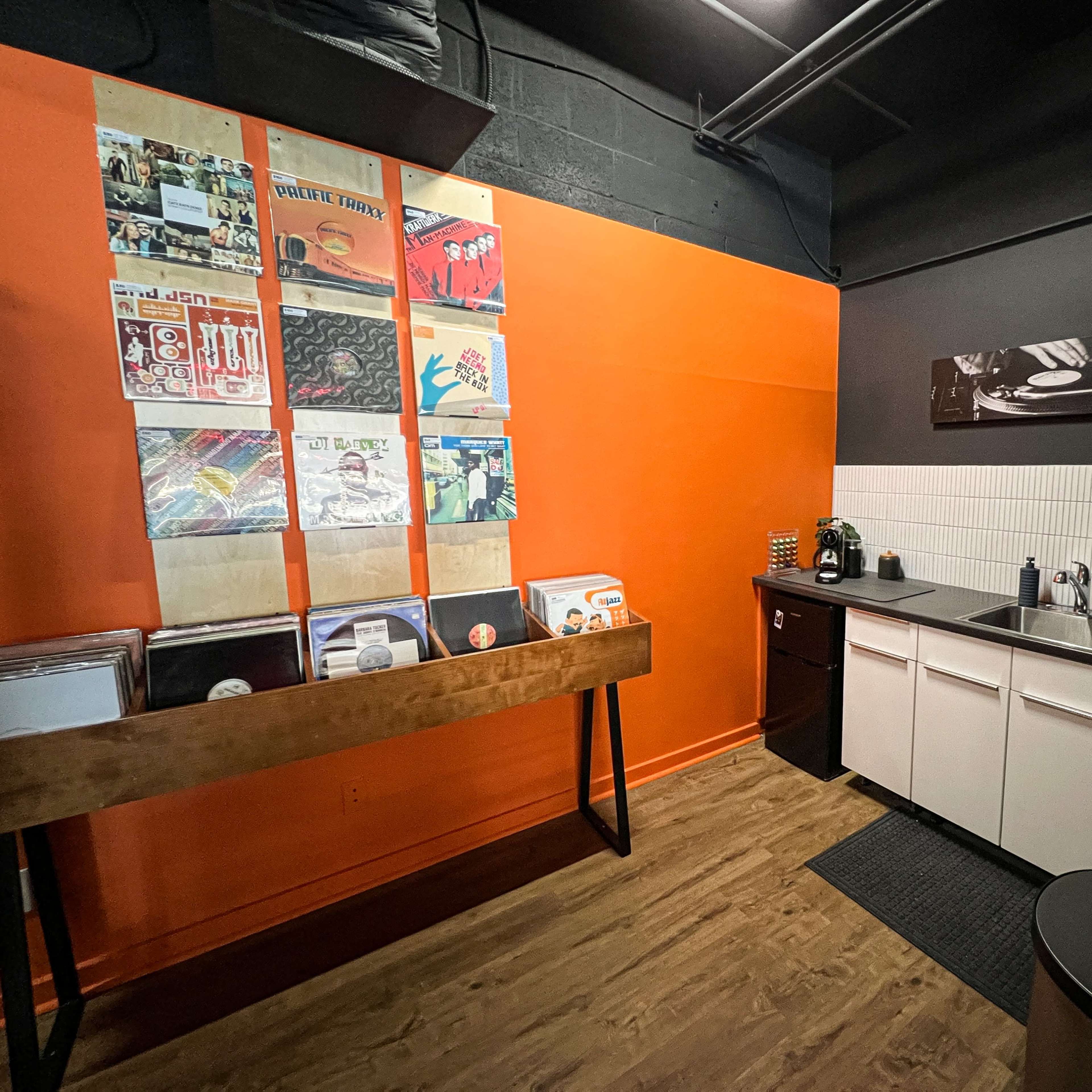 A modern lounge area with a high table surrounded by metal stools, wooden flooring, a wall displaying various artworks, and audio equipment in the background.