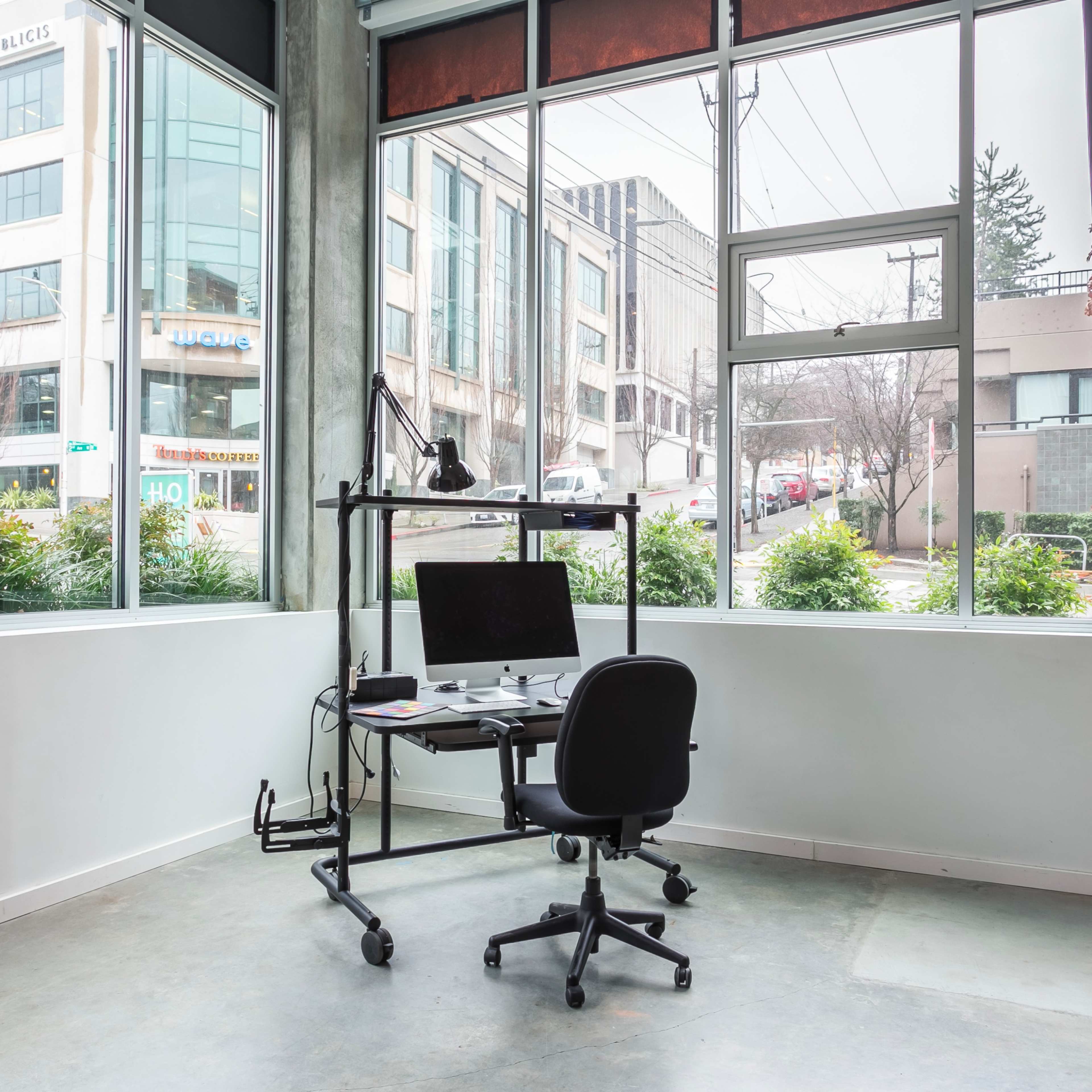 A modern office space with a standing desk and chair positioned near large windows overlooking a city street.