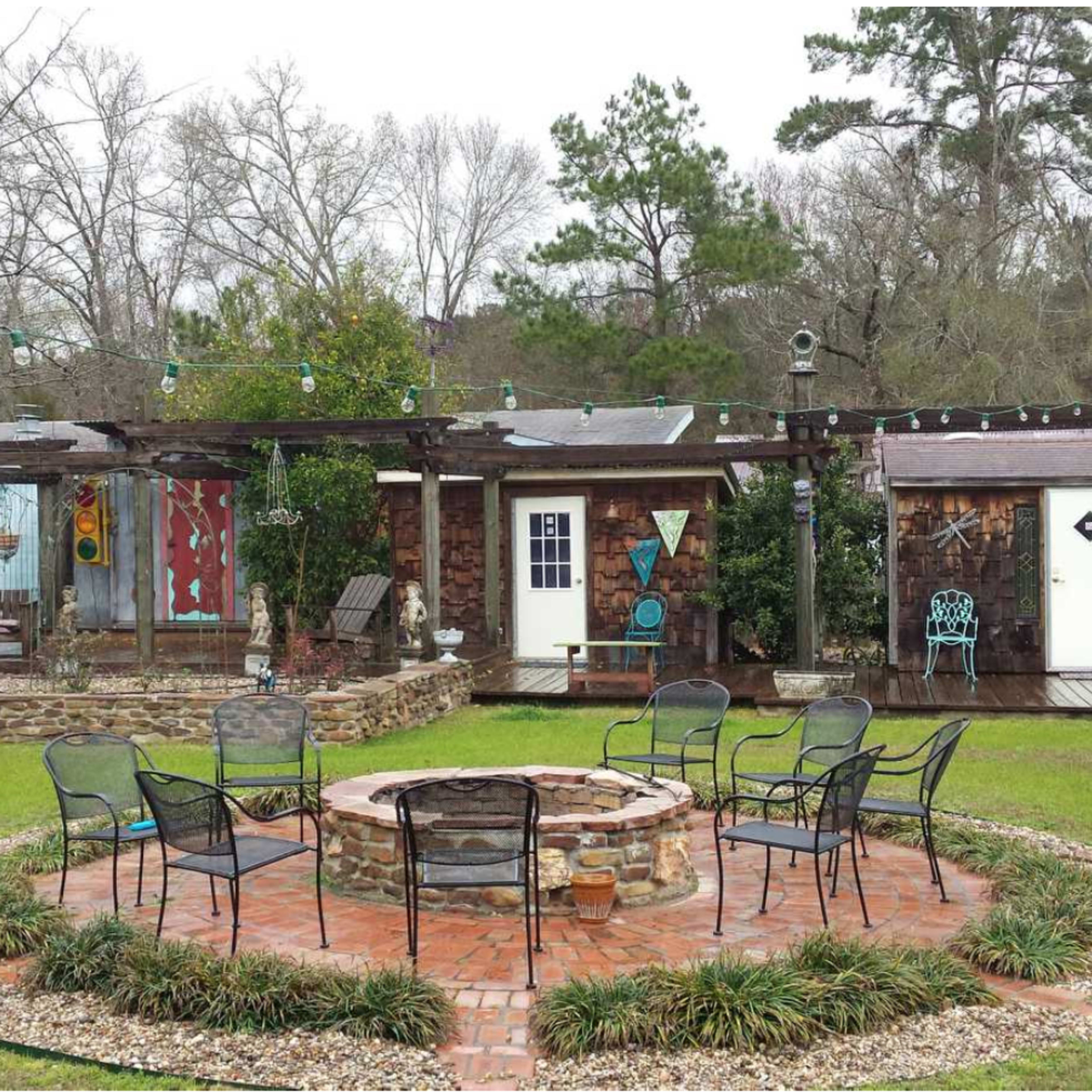 A circular stone fire pit surrounded by several metal chairs in a yard with wooden structures and trees in the background.