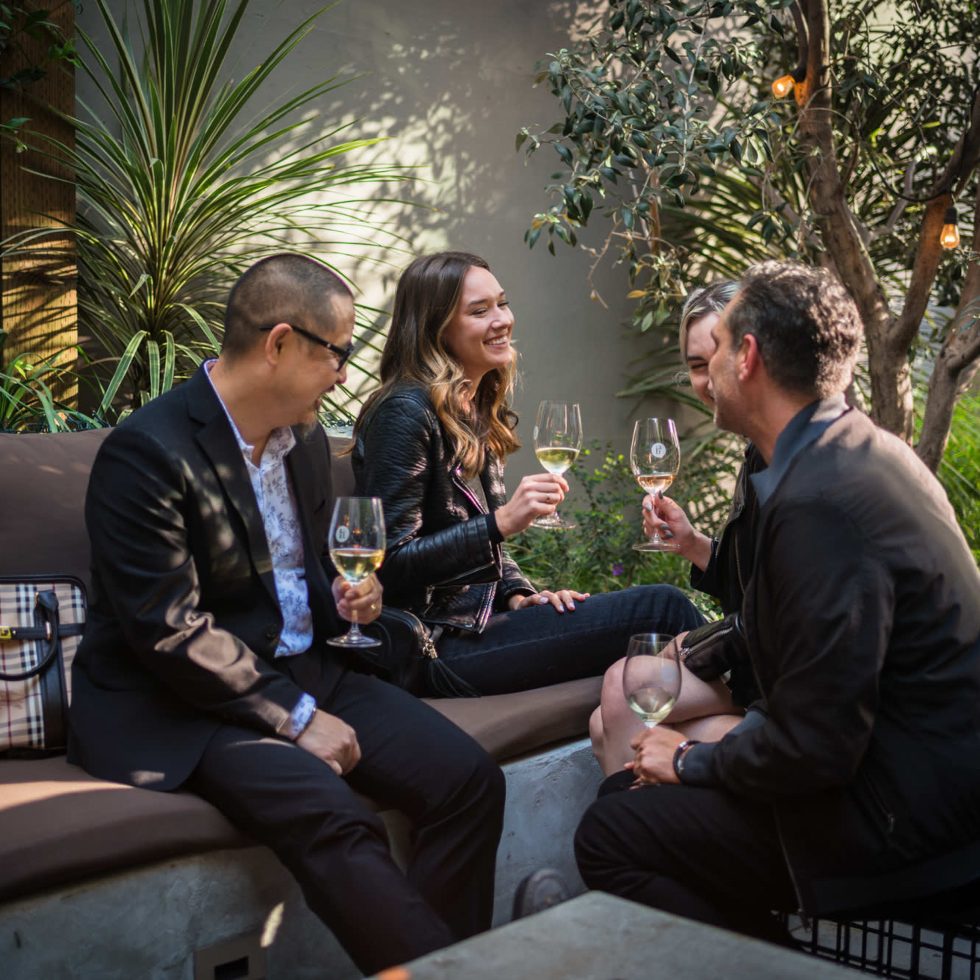 Four people sit together on a bench in a garden setting, enjoying drinks and engaging in conversation.