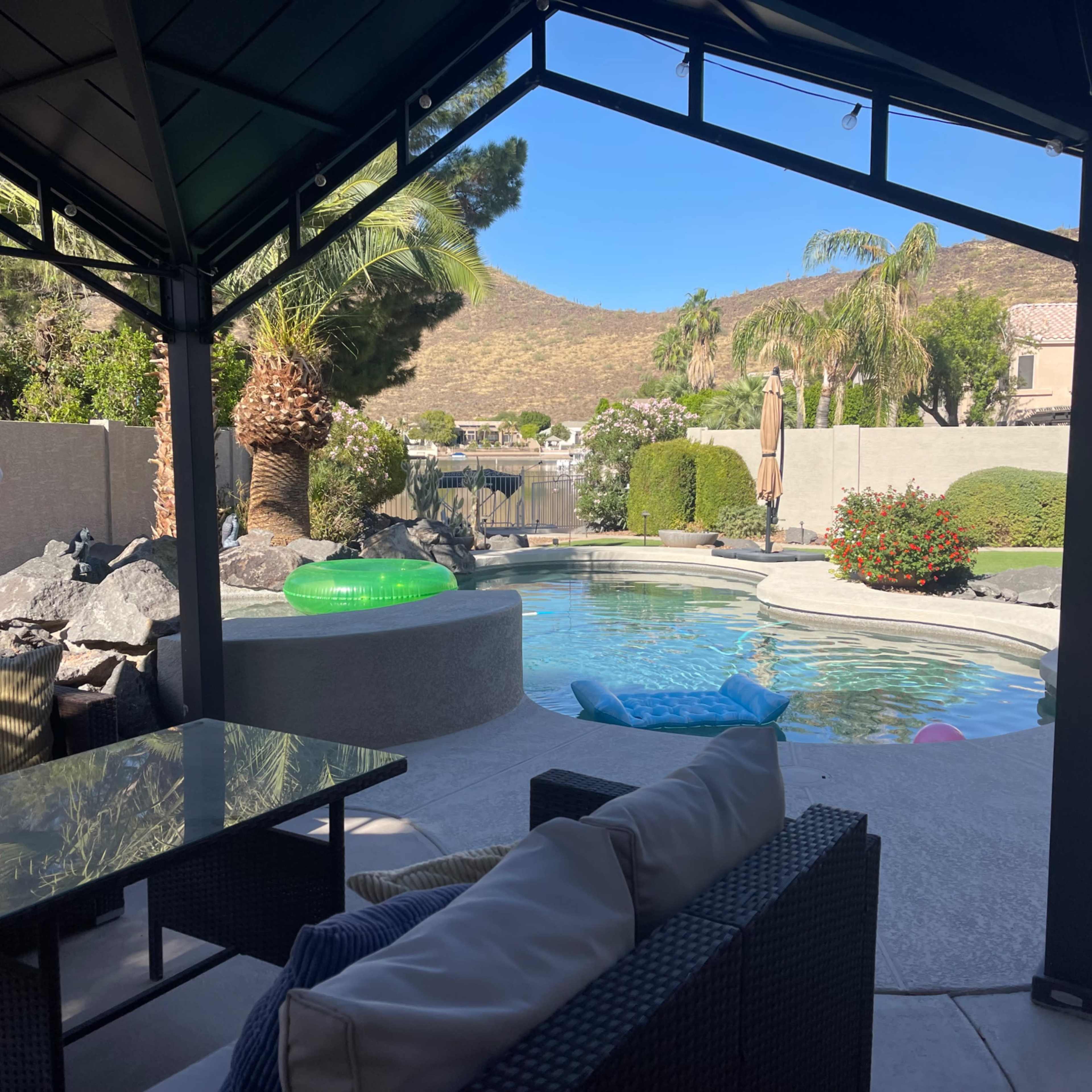 A shaded patio area overlooking a swimming pool surrounded by rocks, greenery, and a scenic view of a hillside.