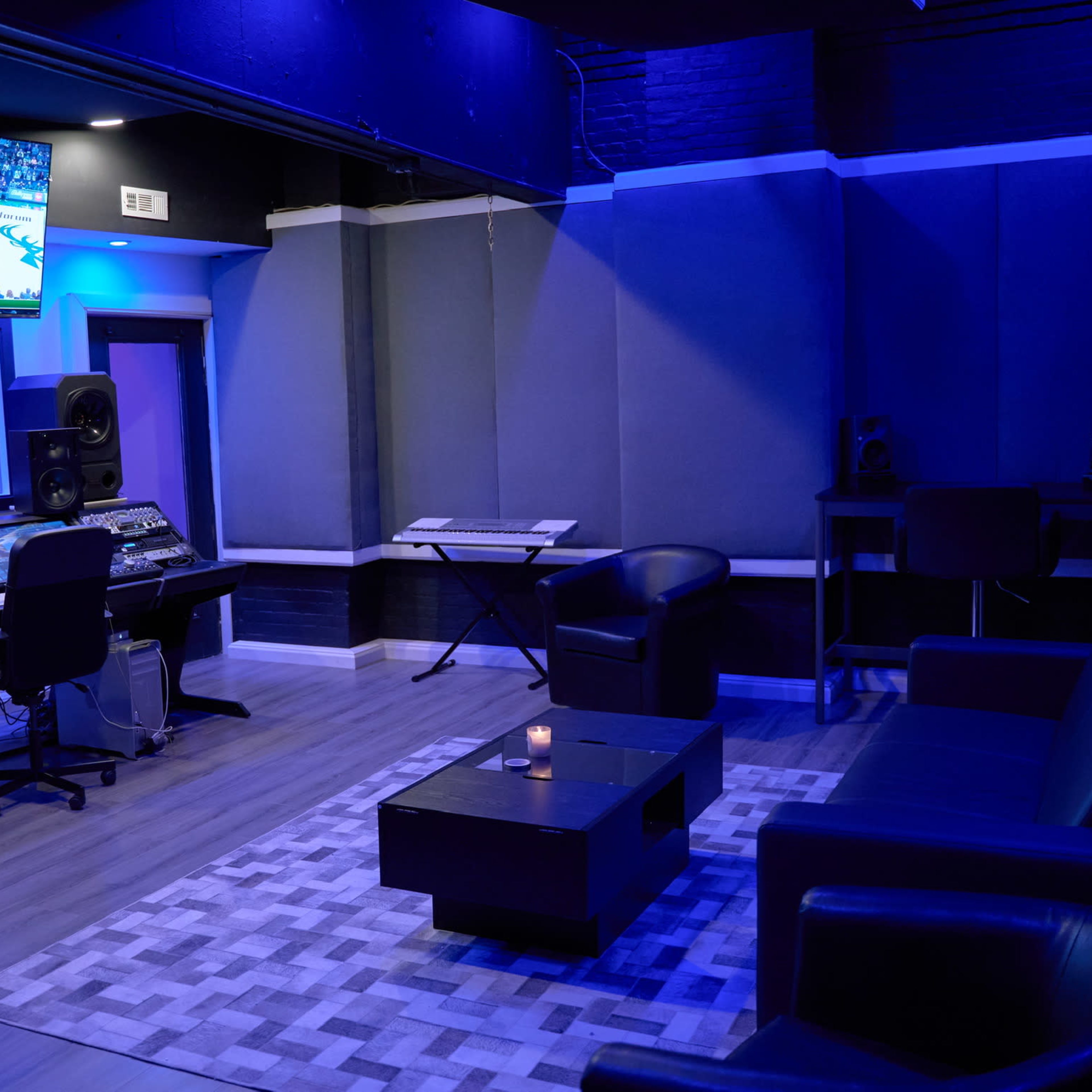 A recording studio is dimly lit in blue tones, featuring a central control desk, two black chairs, and a TV mounted on the wall displaying a sports game.