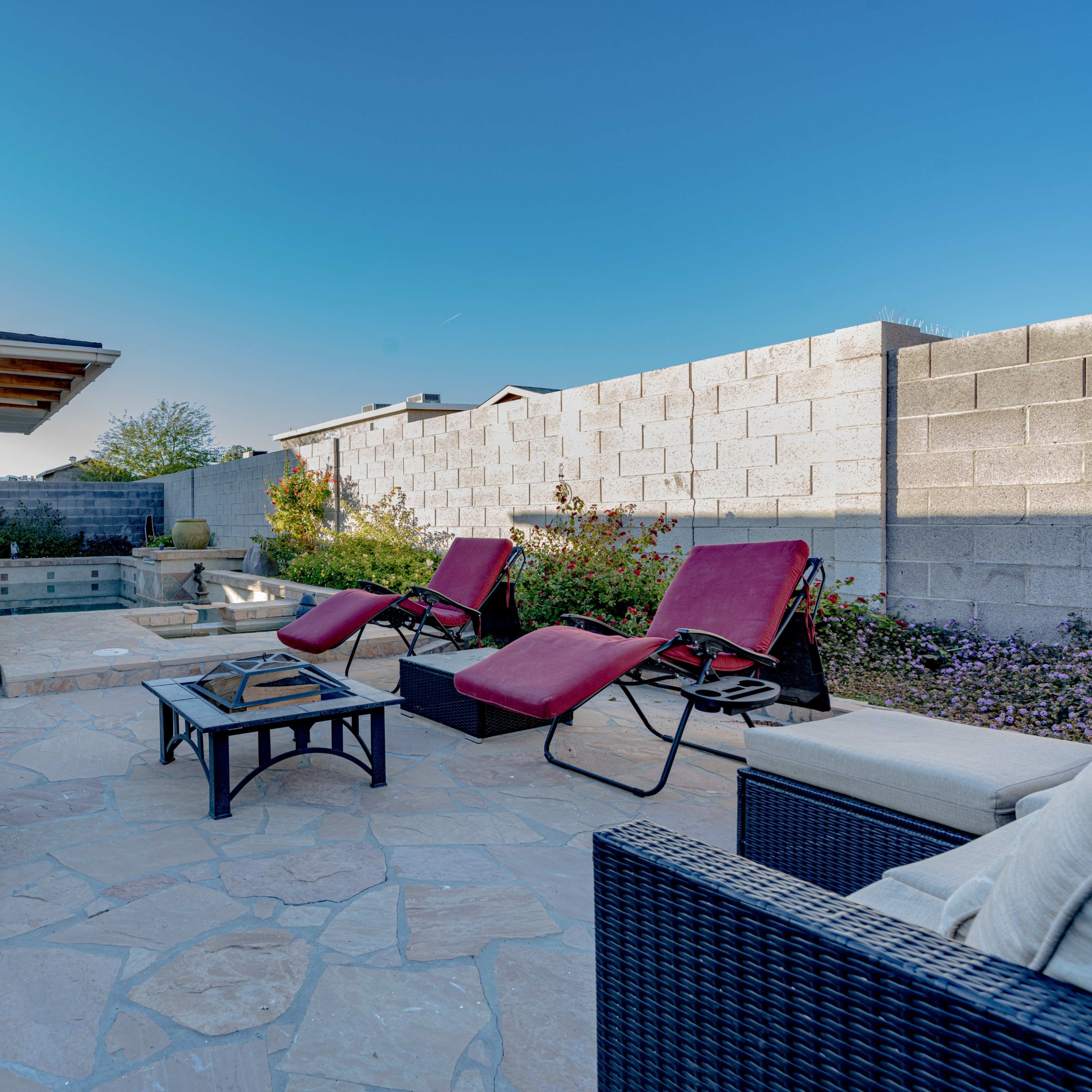 A backyard patio features red lounge chairs, a small table, and a hot tub surrounded by a stone wall and vegetation.
