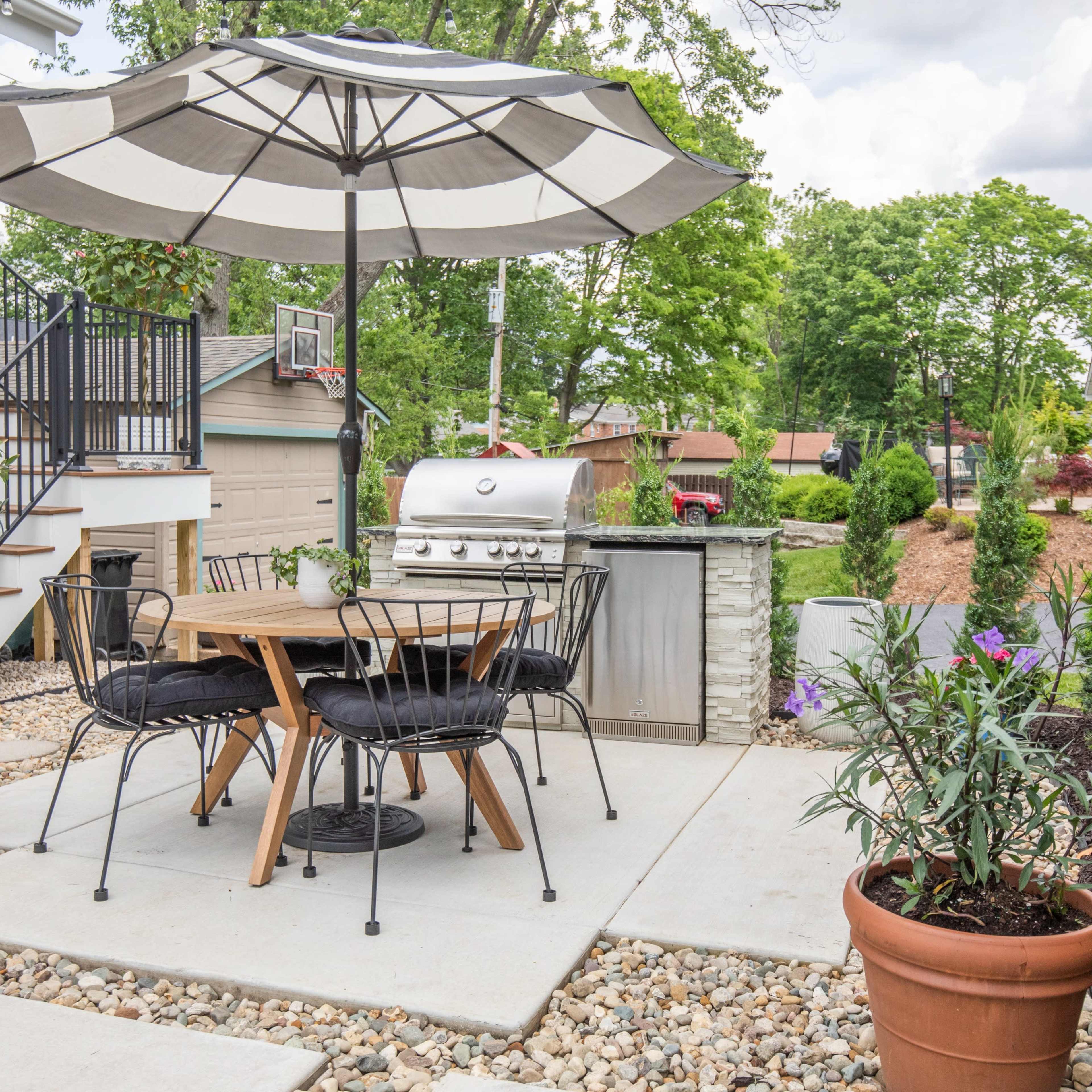 A patio area features a table with chairs under a large umbrella, a grill nearby, and landscaped greenery surrounding the space.