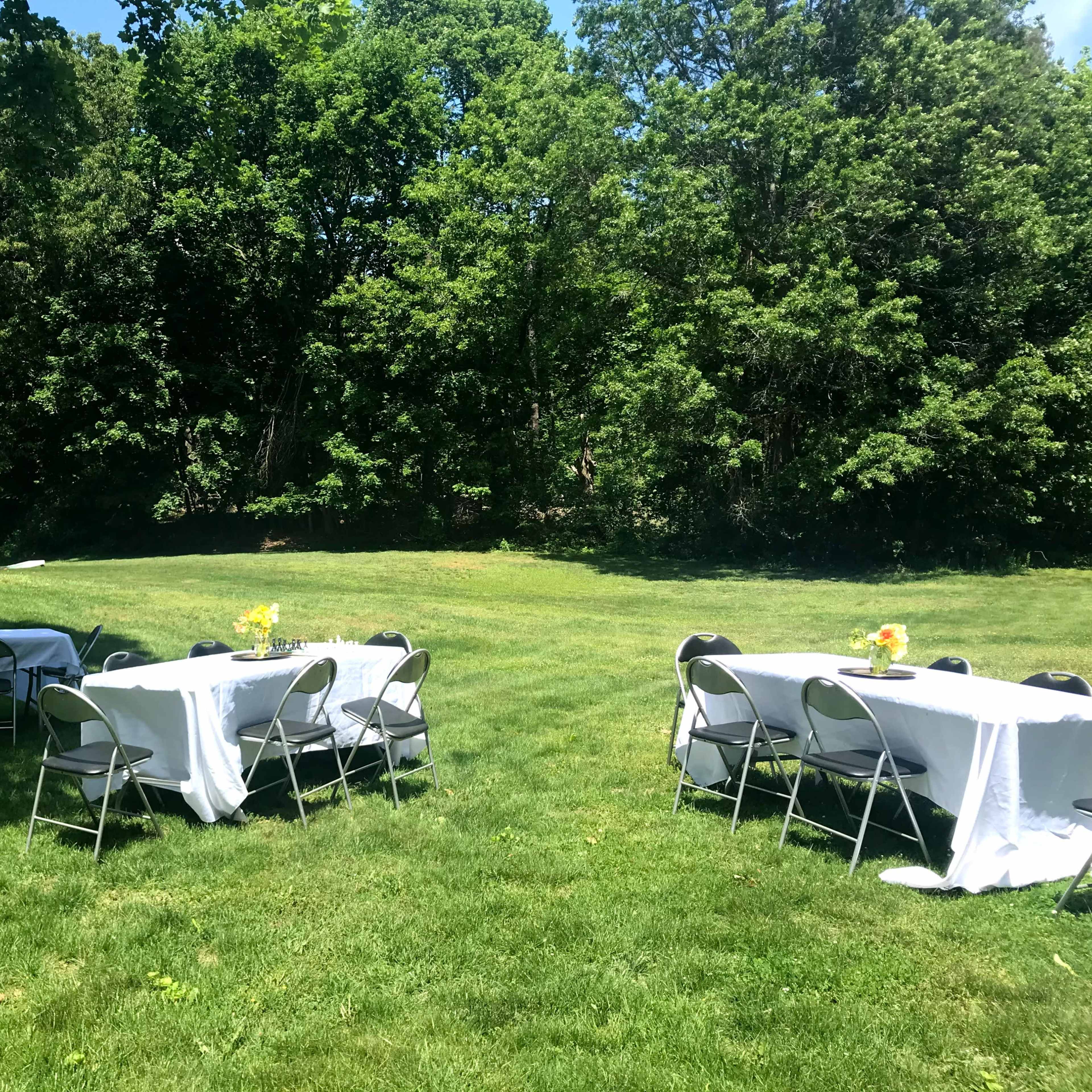 Several tables with white tablecloths set up on a green lawn surrounded by trees.