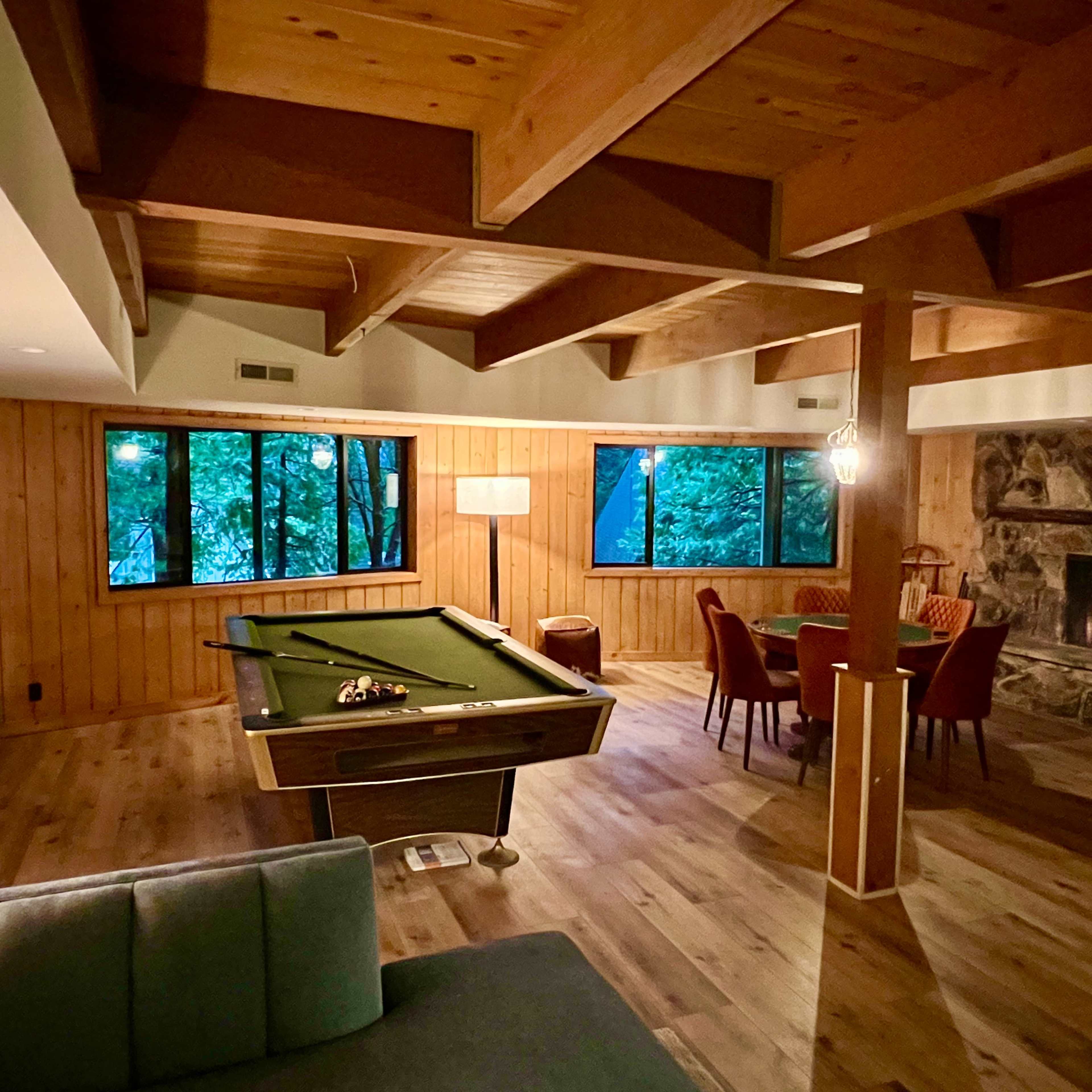 A spacious, wooden-paneled living room features a billiard table, a dining area, and large windows overlooking a wooded landscape.