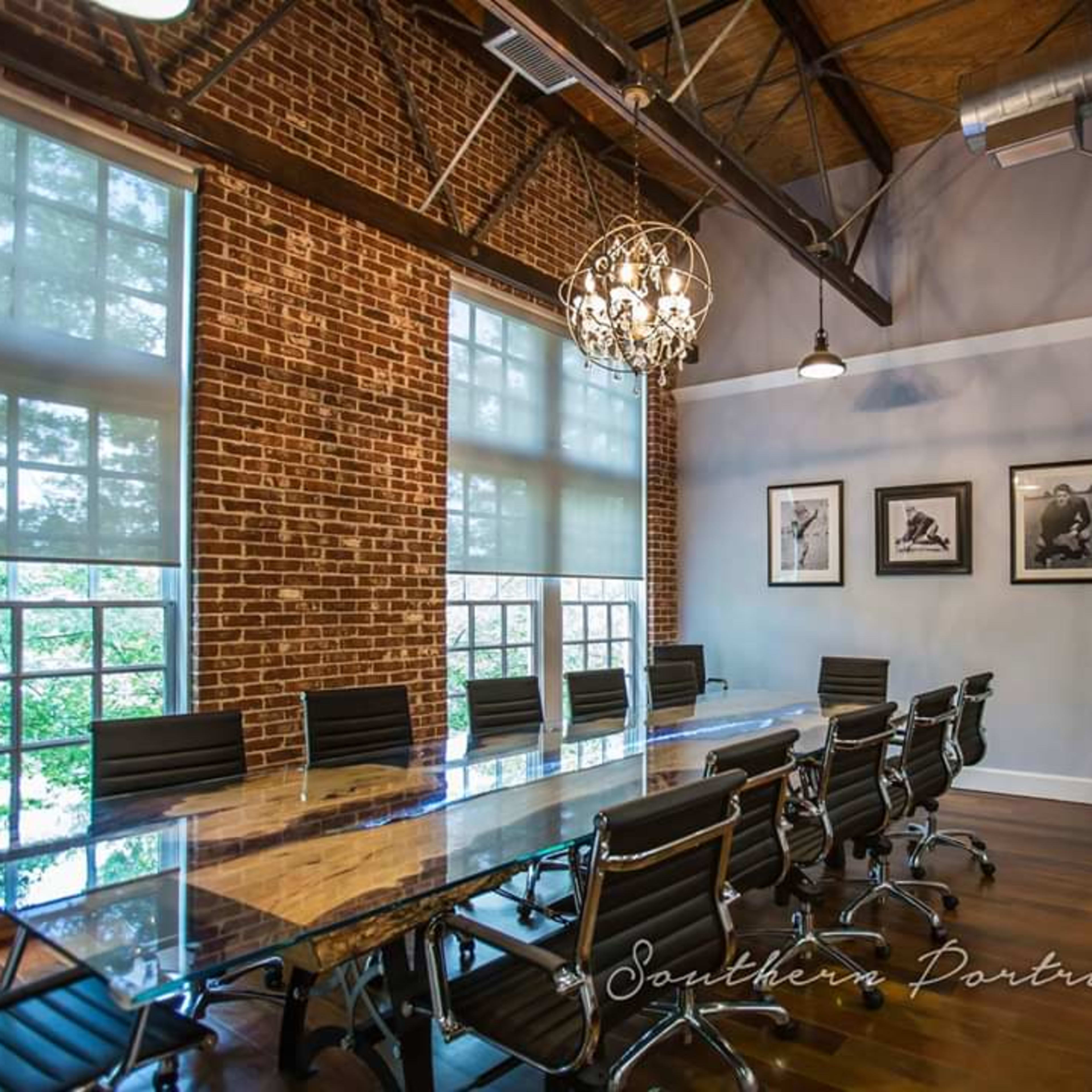 A modern conference room featuring a long glass table, black office chairs, exposed brick walls, and framed artwork on the walls.
