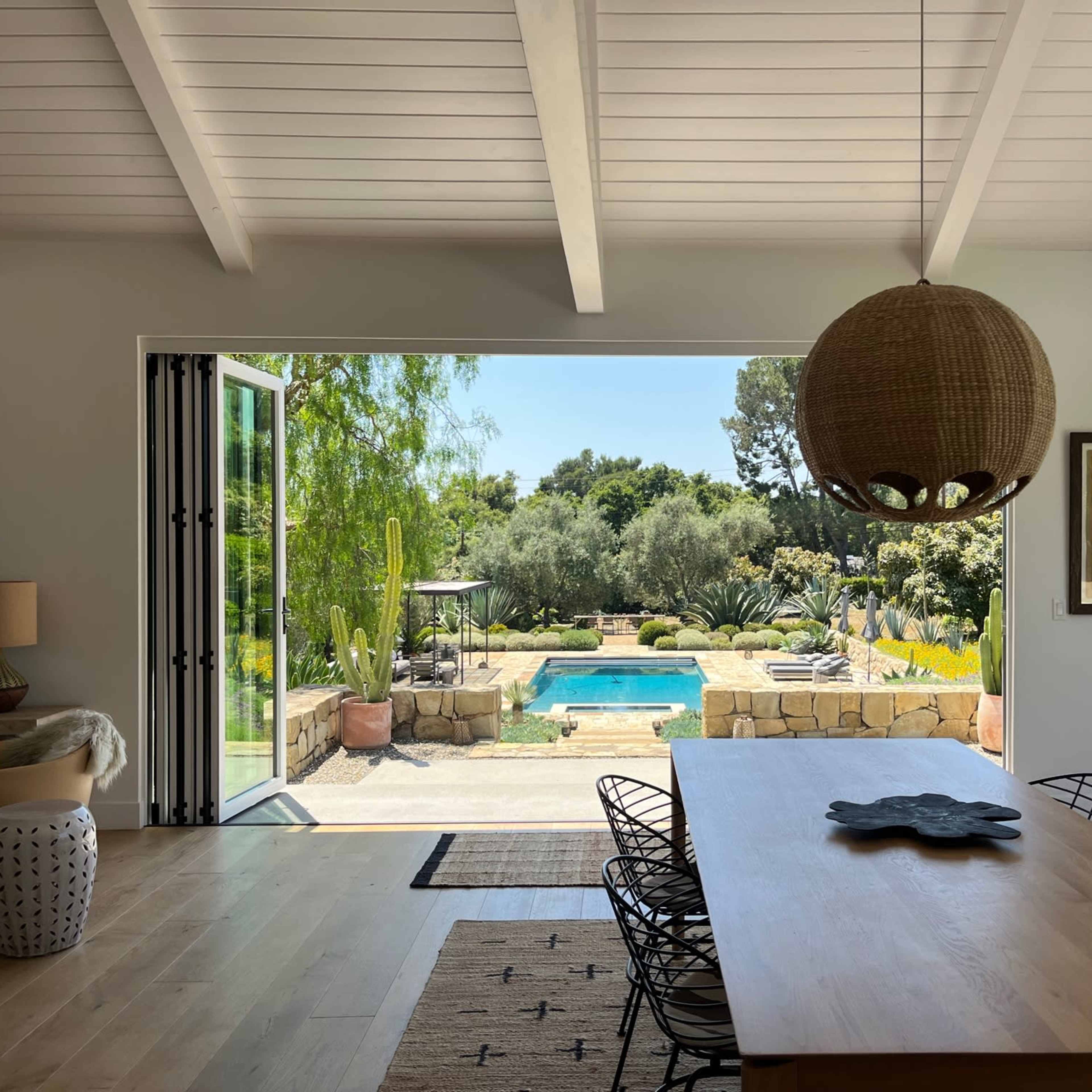 A dining area features a large table and opens to a backyard with a pool surrounded by greenery and landscape elements.