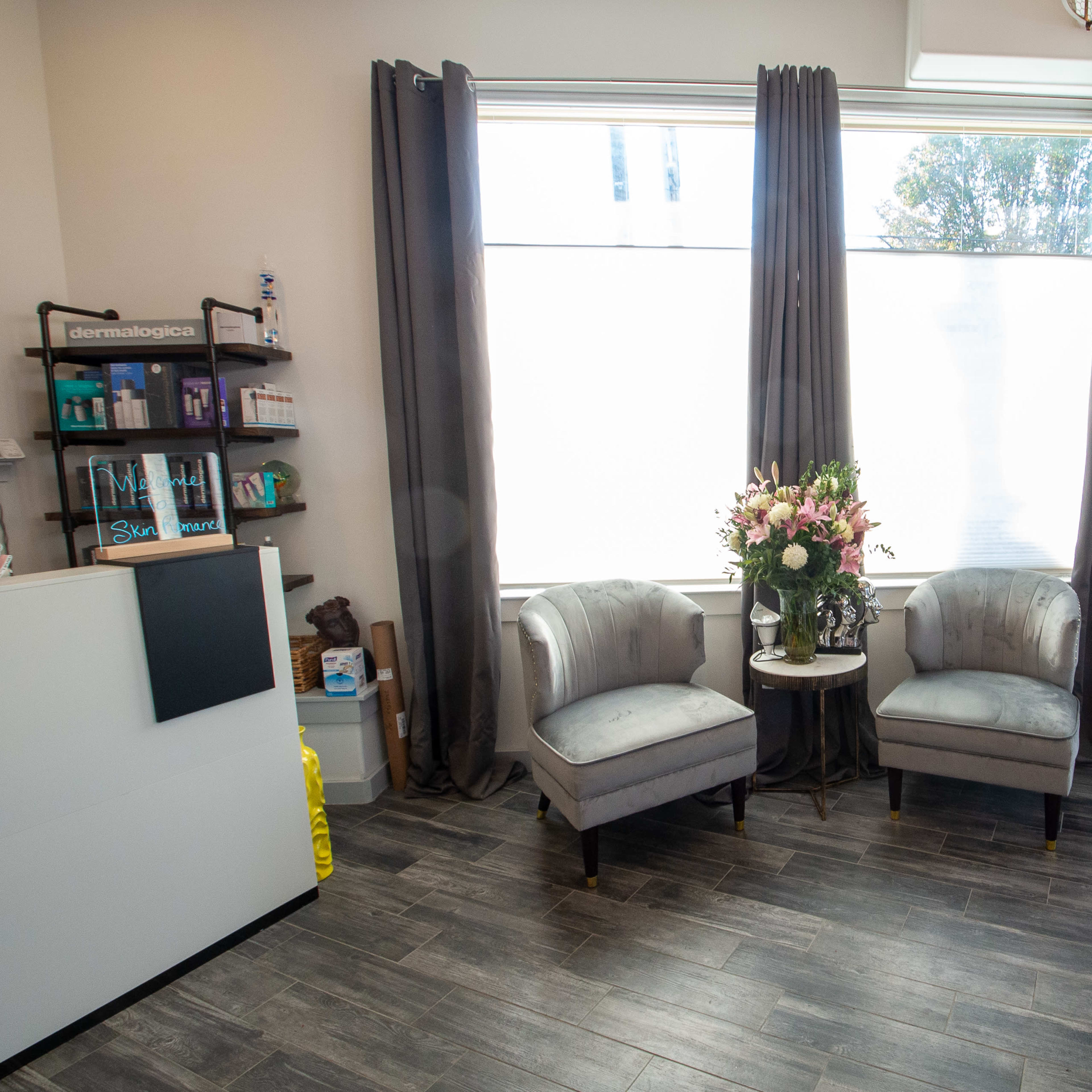 A skincare clinic reception area with two gray chairs and a small table decorated with a flower arrangement, alongside a bookshelf and a wall-mounted water feature.
