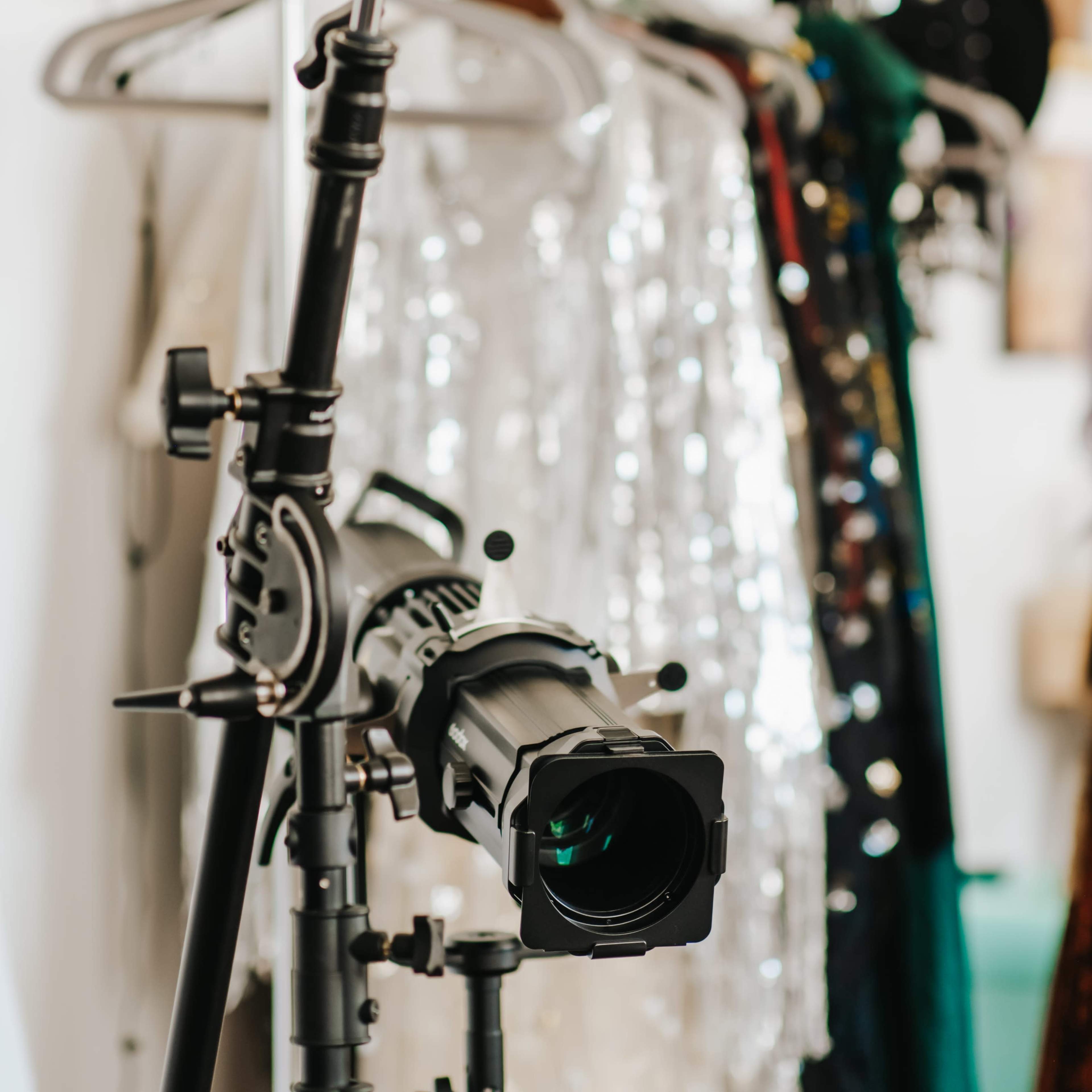A film lighting setup with a spotlight and camera lens is positioned in front of racks of sequined clothing.