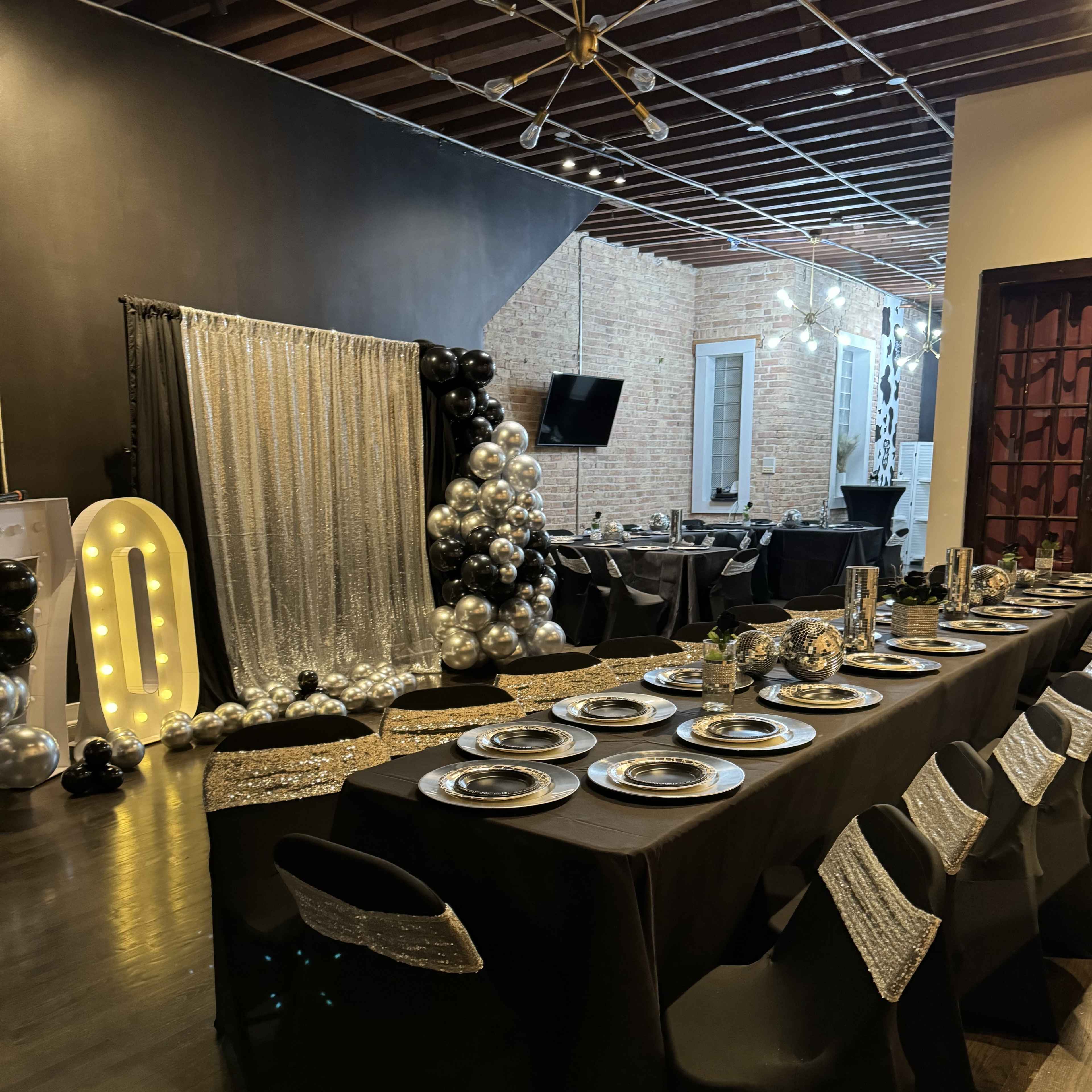 A decorated dining area with a long black table set with silver plates, surrounded by black chairs and adorned with metallic balloons.
