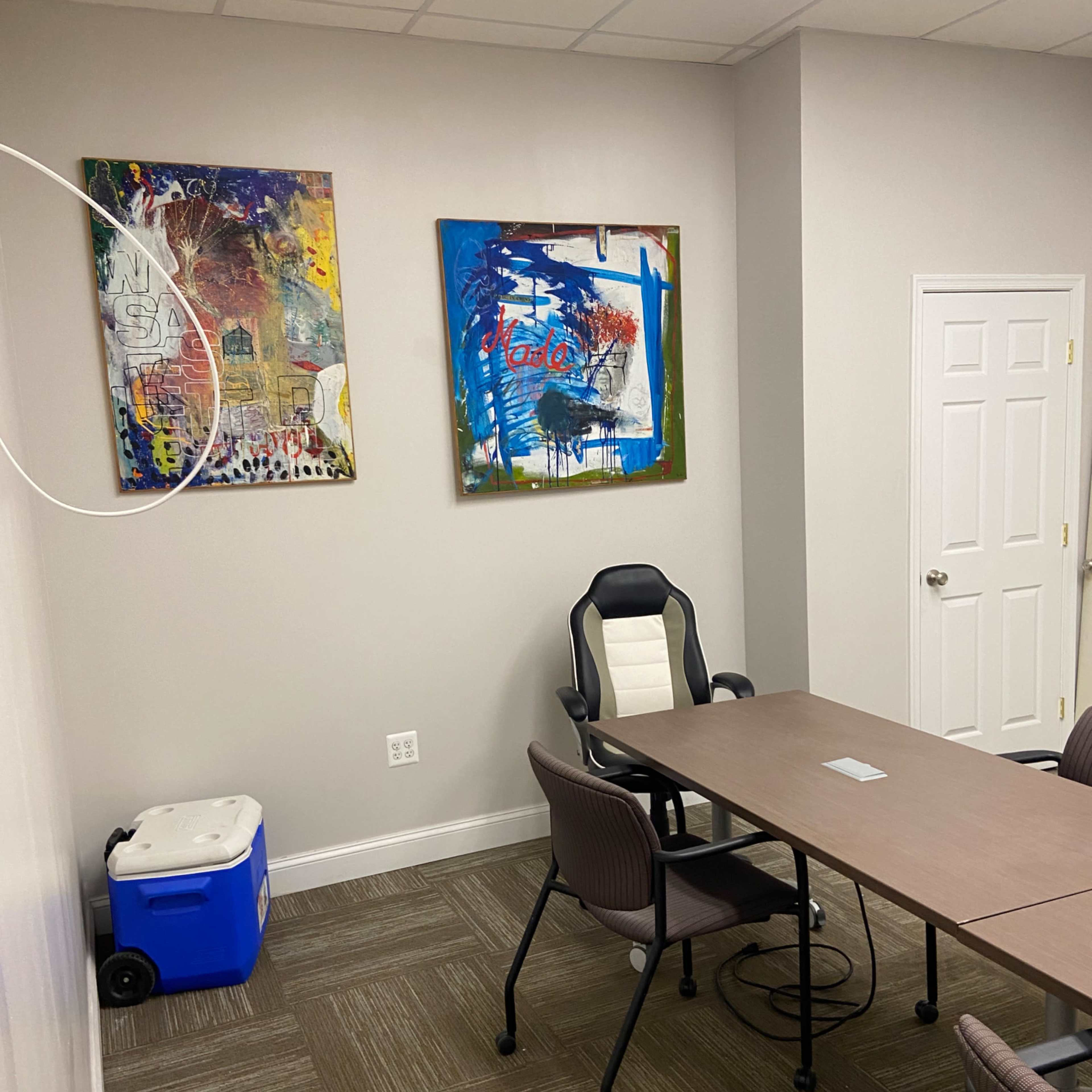 A small meeting room with a rectangular table, several chairs, two colorful paintings on the wall, and a cooler in the corner.