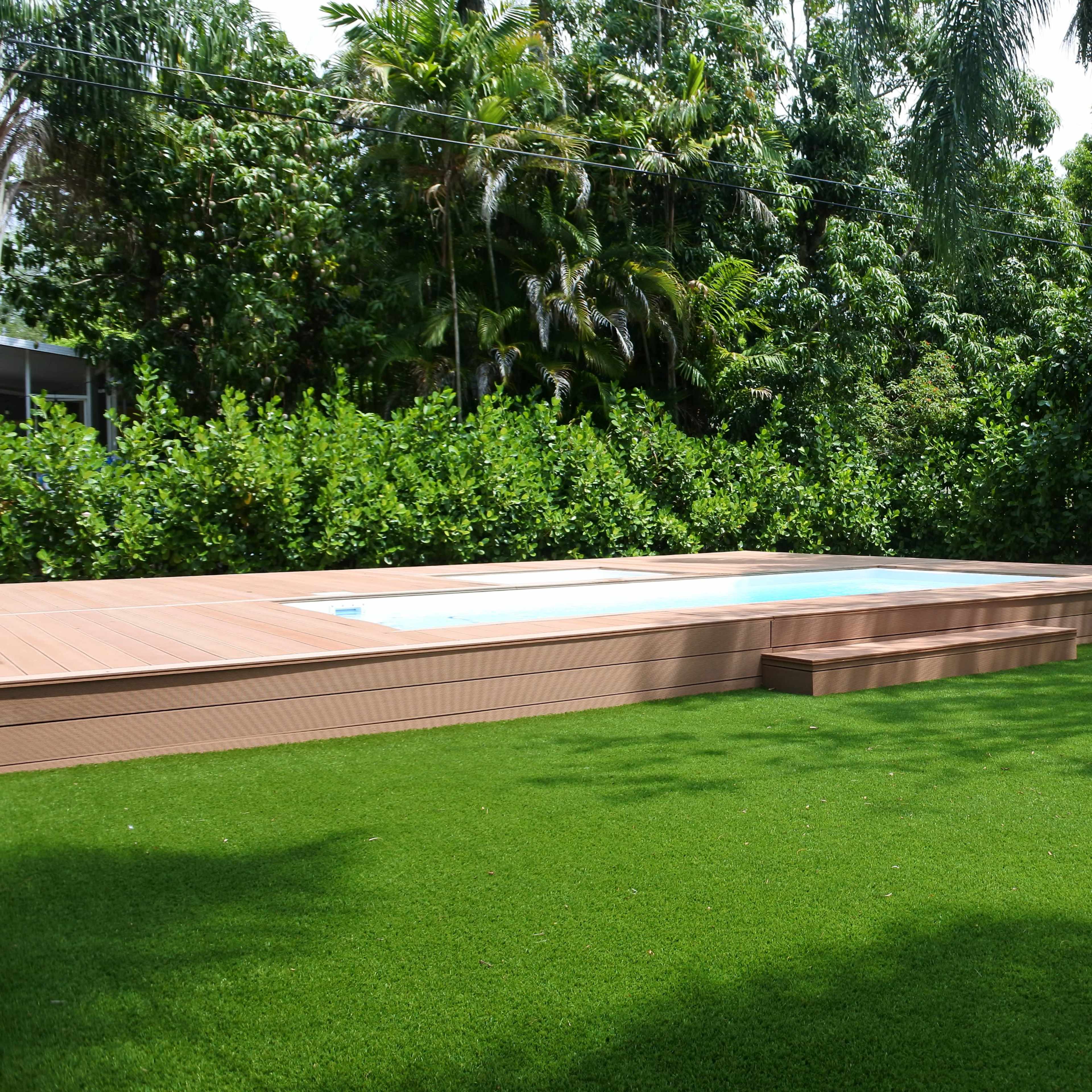 A wooden deck surrounds a small swimming pool set in a grassy area with lush greenery in the background.