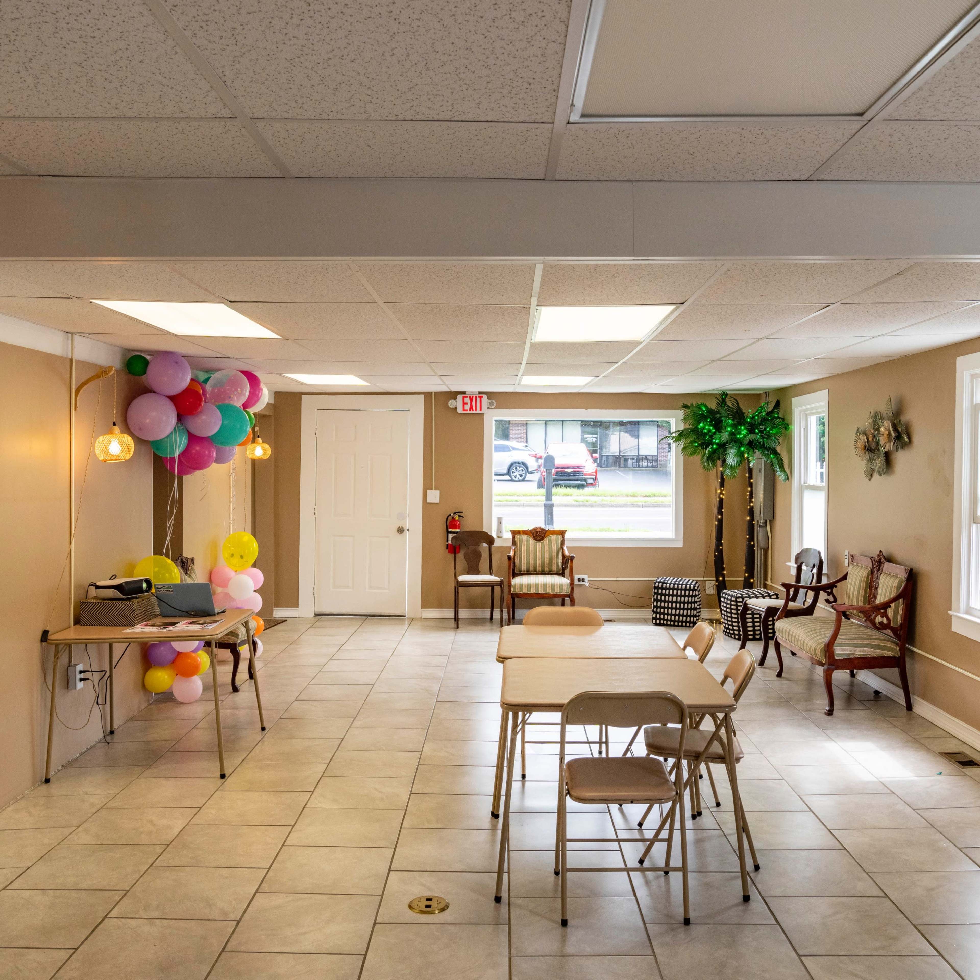 A spacious, well-lit indoor area with tables and chairs arranged, balloons attached to a wall, and a projector set up on a table.