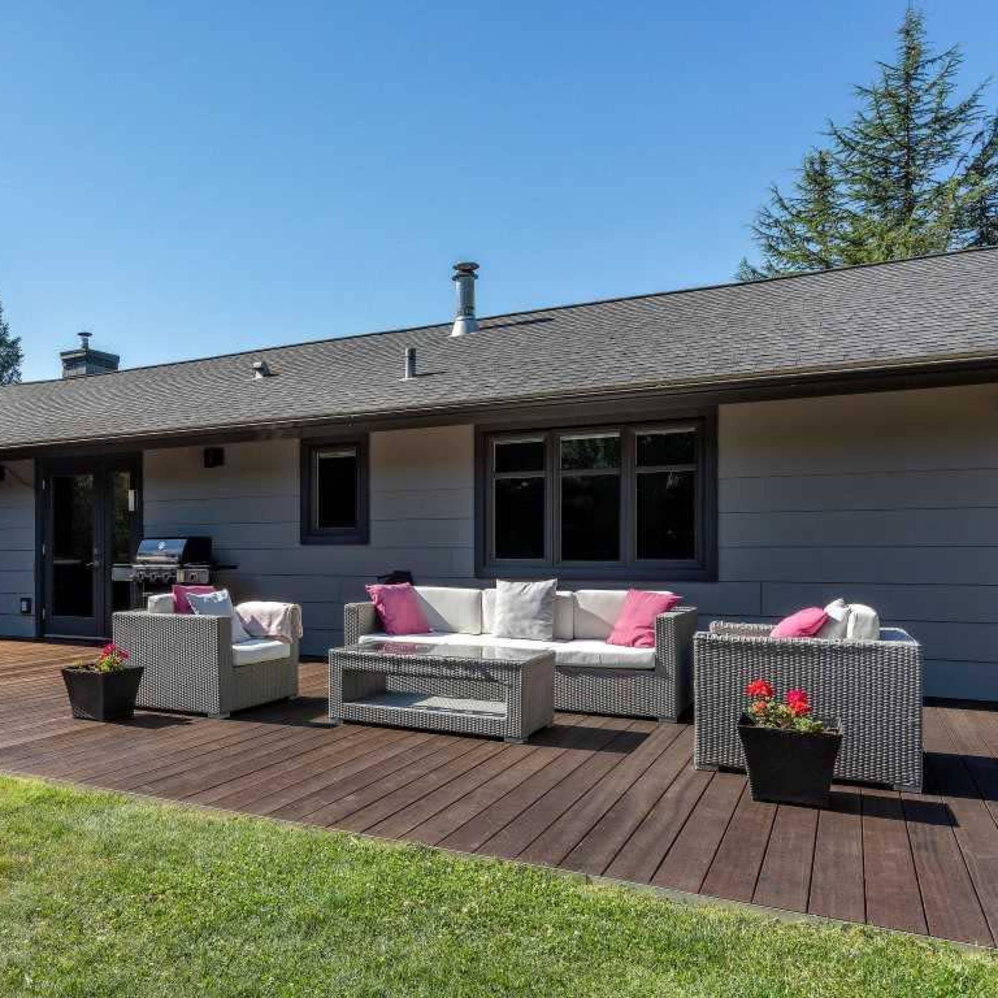 A backyard patio with a wooden deck, featuring outdoor seating and a dining area surrounded by grass and trees.