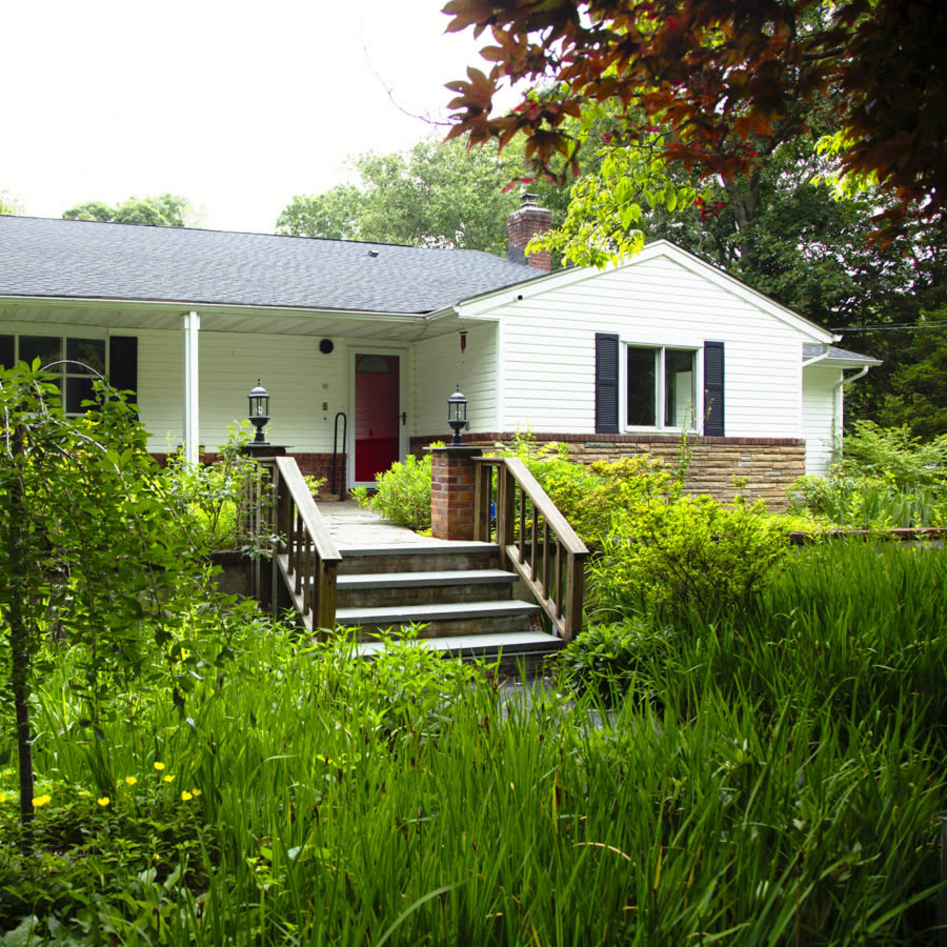 A single-story white house with a red door is surrounded by lush greenery and a set of stairs leading to the front porch.