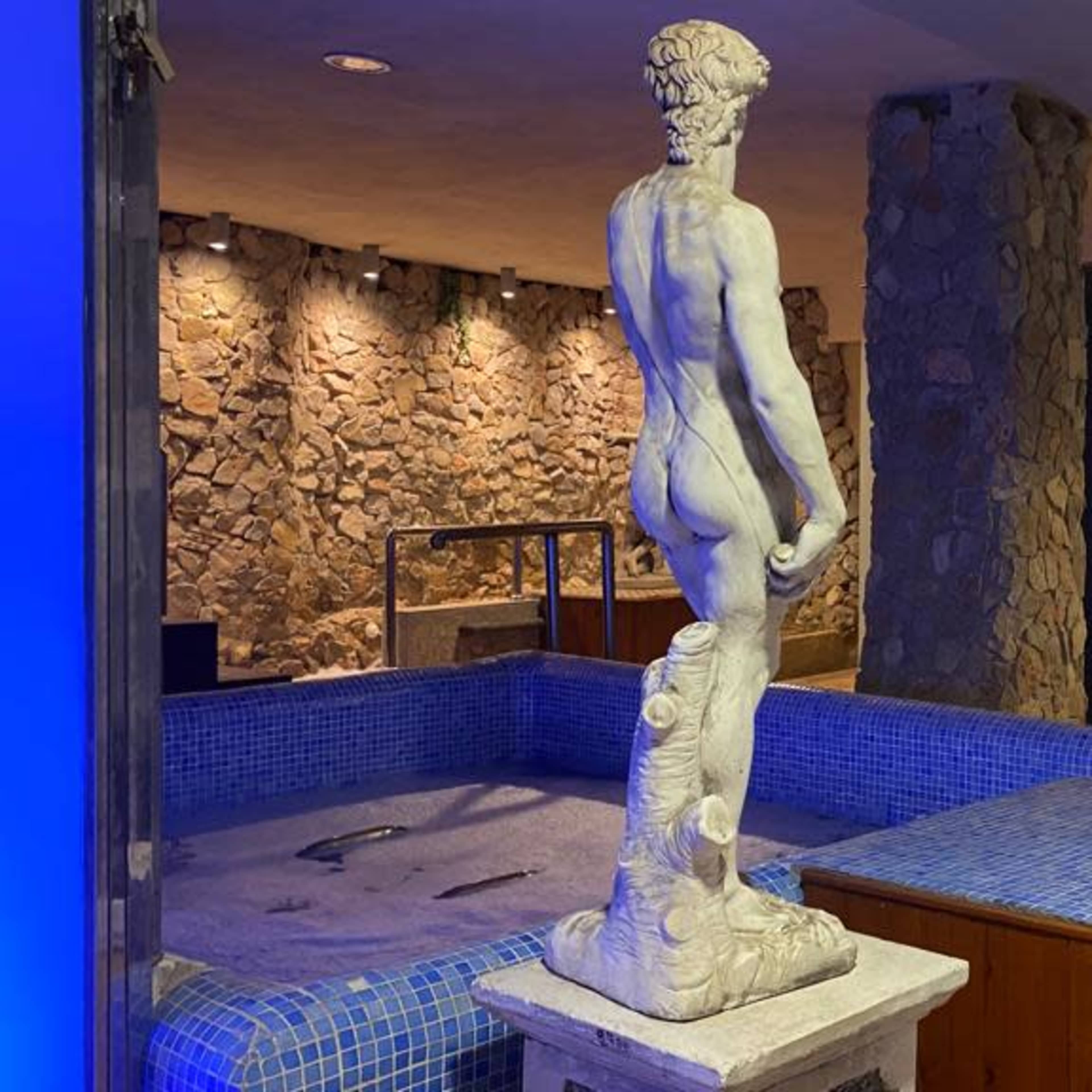 A marble statue of a nude male figure stands beside a blue-tiled hot tub in a spa-like setting with stone walls.