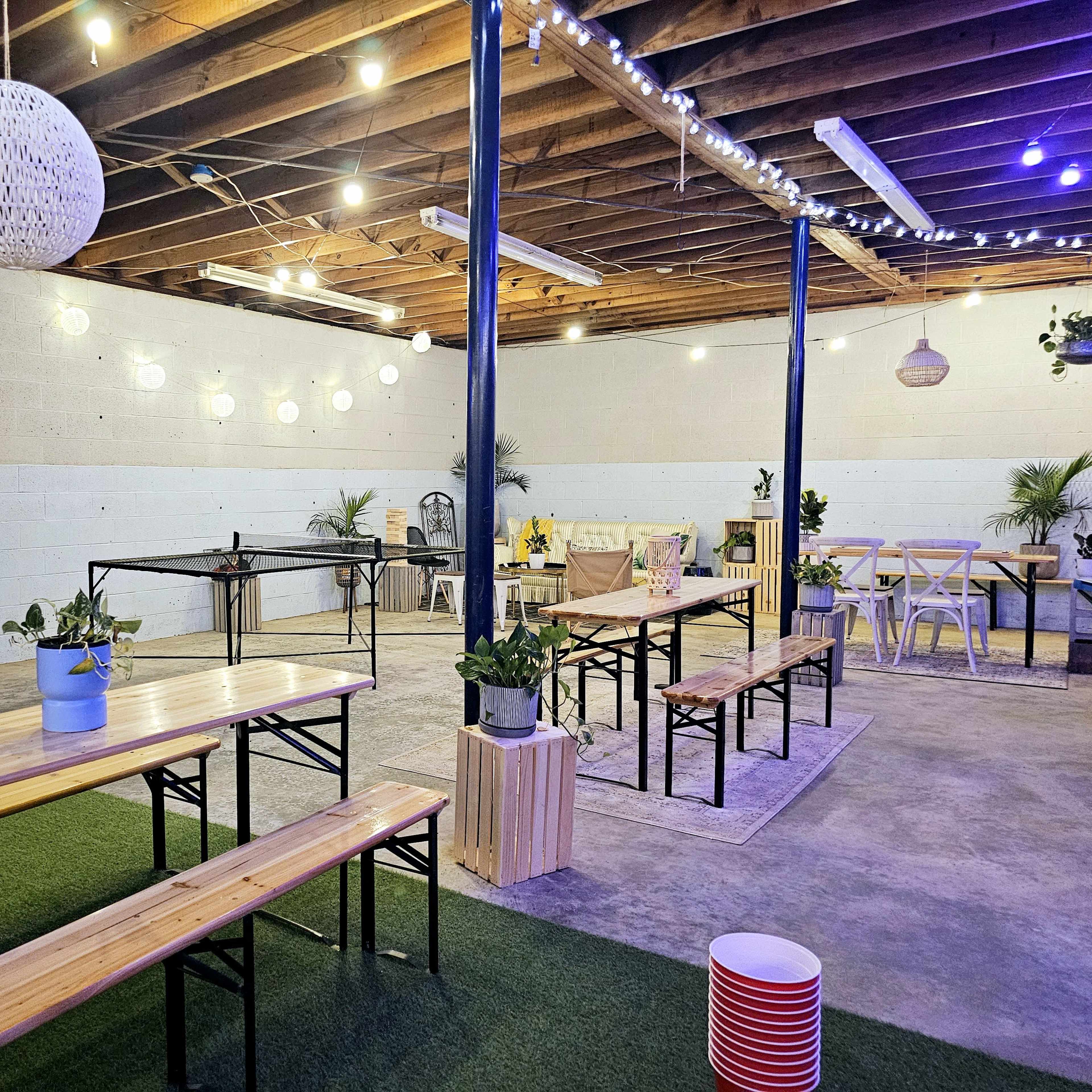 An indoor space with wooden tables and benches, green plants, and decorative lighting hanging from the ceiling.