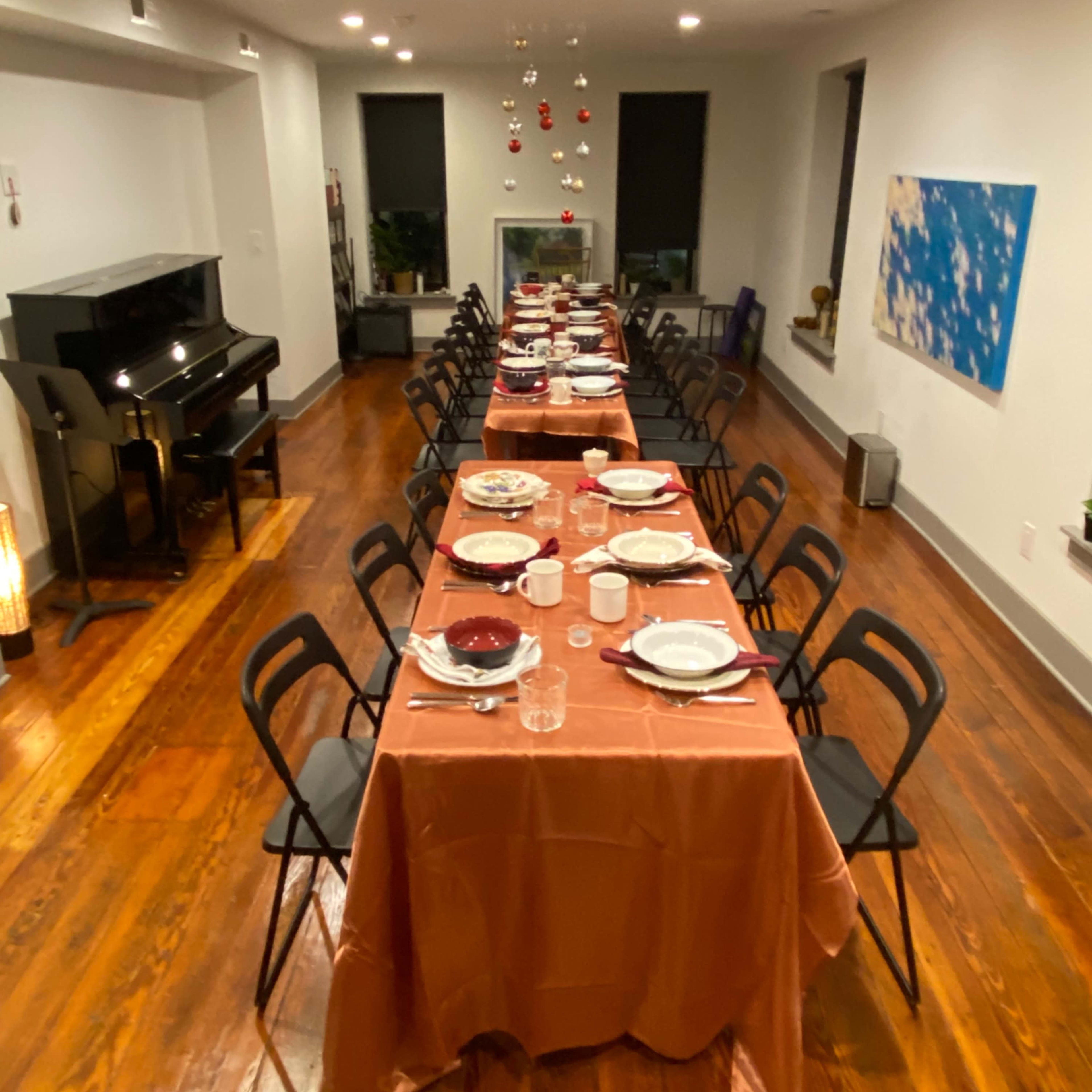 A large dining room features two long tables set with dinnerware and decorations, illuminated by overhead lights, with a piano in the corner.