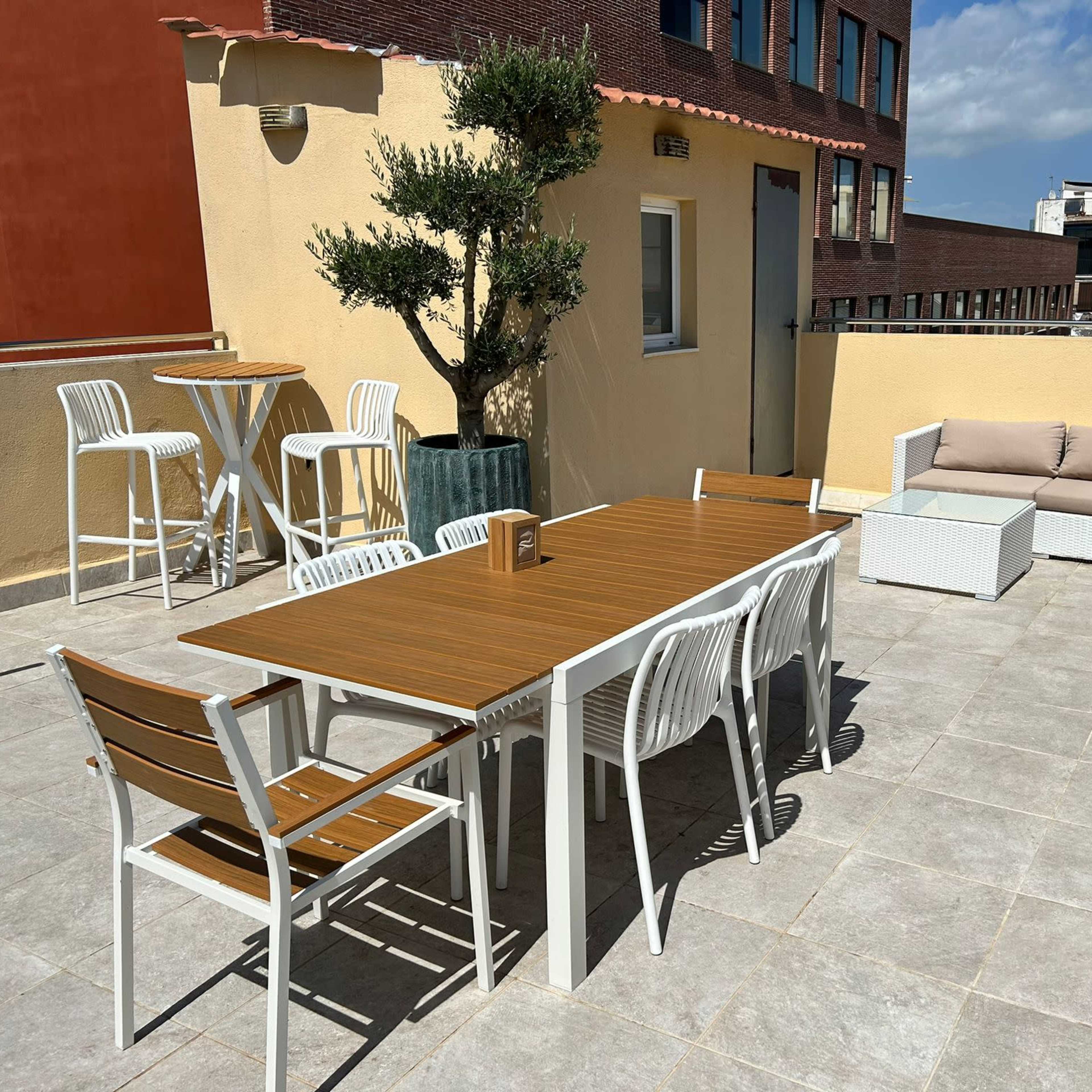 A rooftop terrace featuring a wooden table with several chairs, a couch, potted plants, and a view of surrounding buildings under a clear sky.