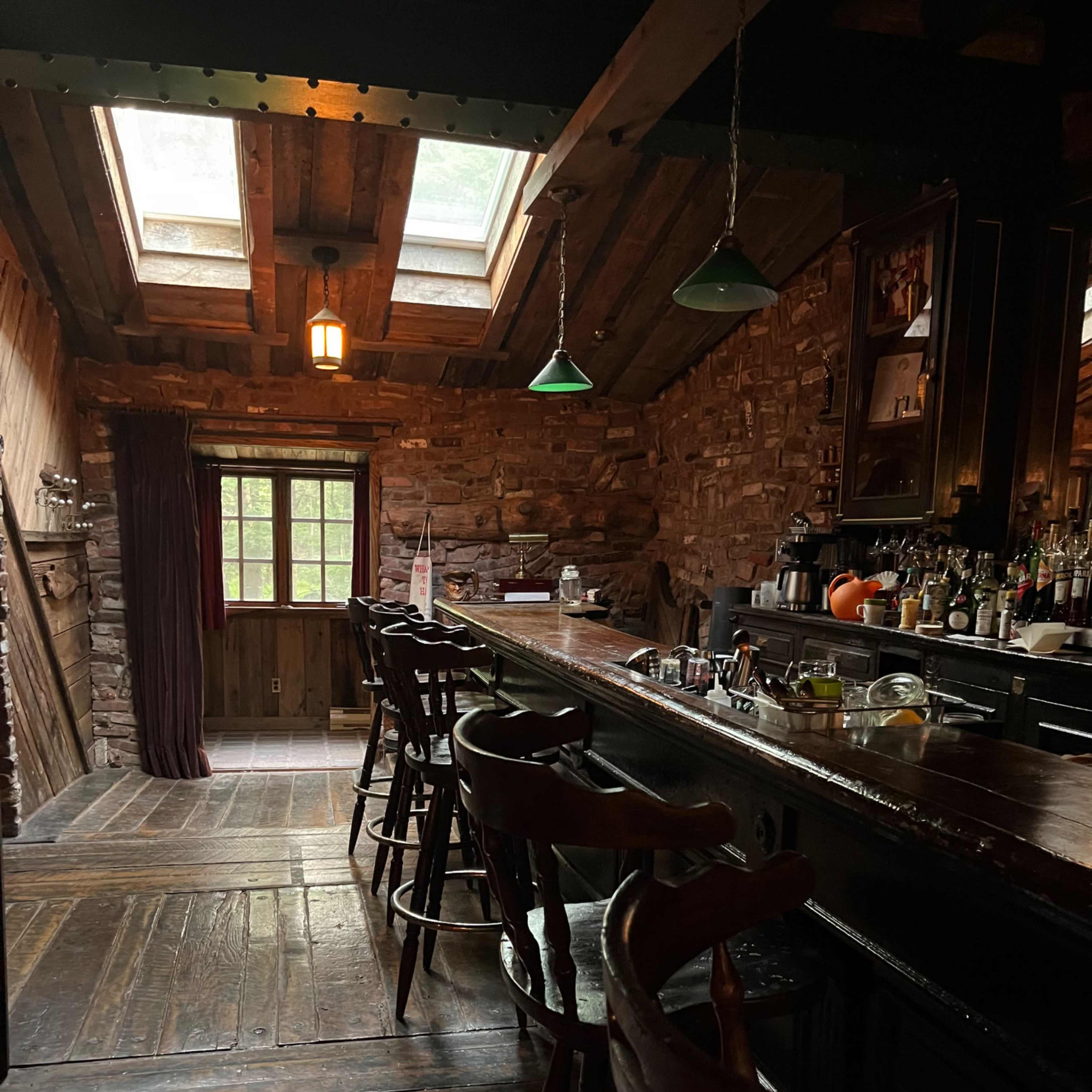 A rustic bar area with a wooden countertop, high stools, and a stone wall, illuminated by natural light from skylights above.