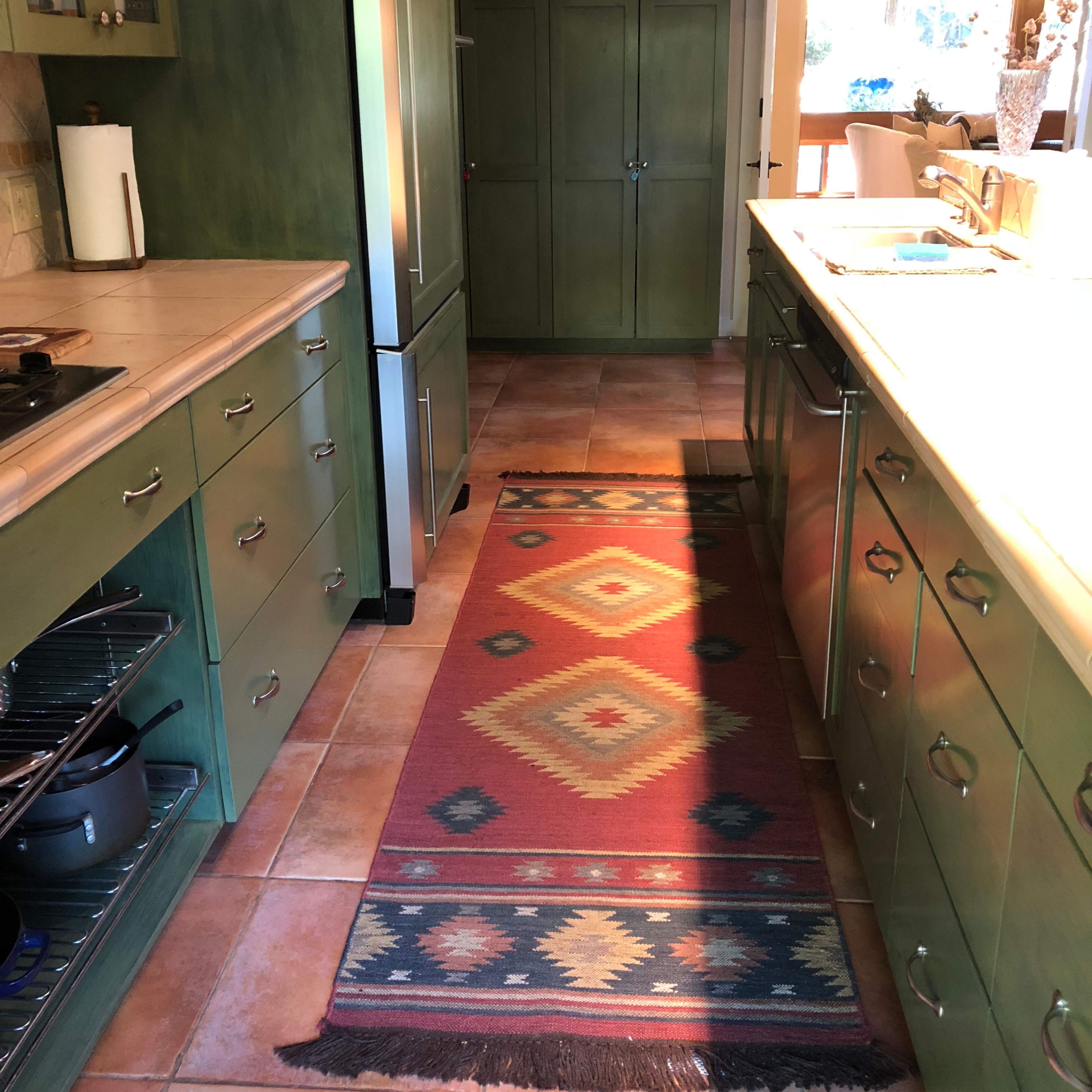 A kitchen with green cabinets, a patterned rug running the length of the floor, and an open layout leading to a dining area.
