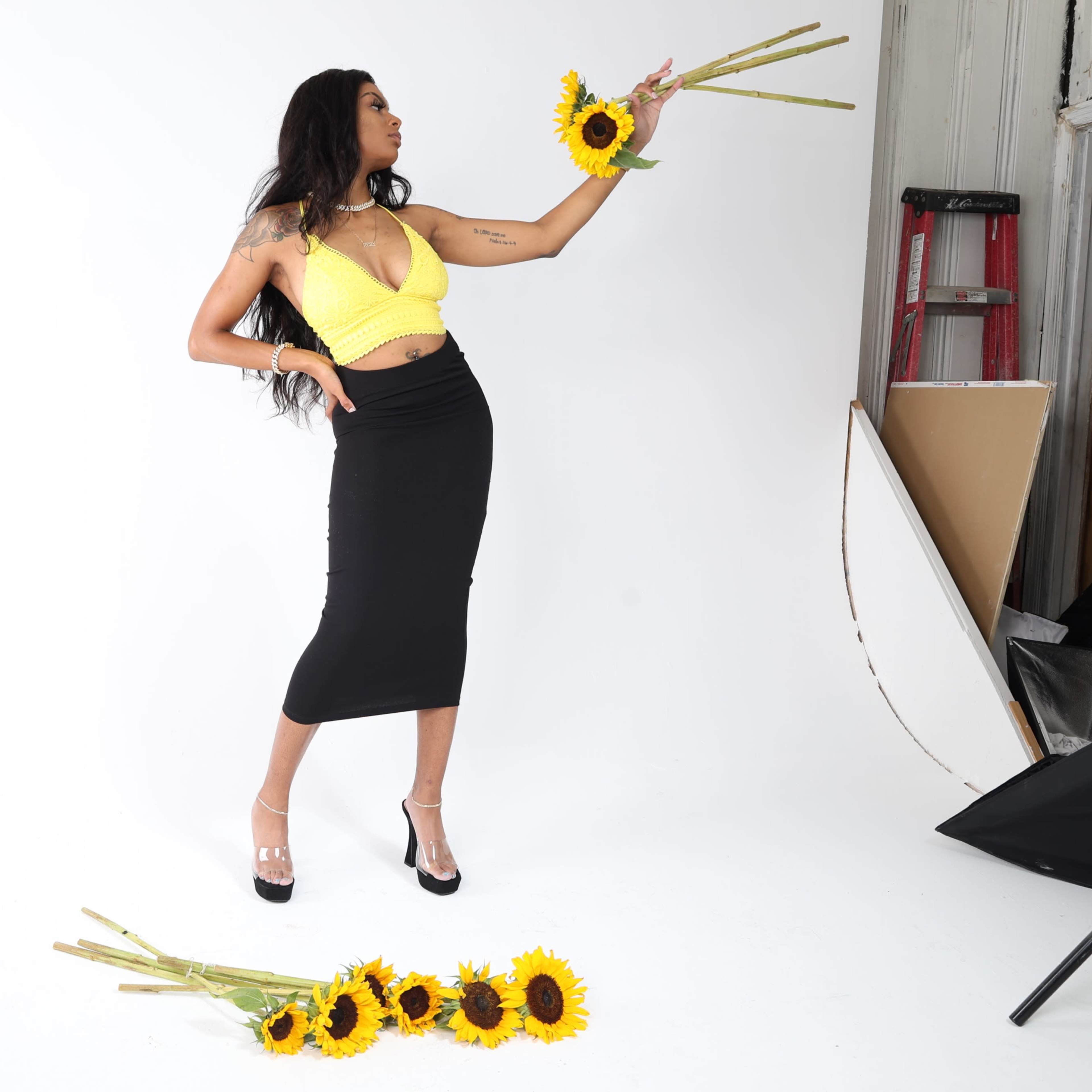 A woman in a yellow top and black skirt poses with sunflowers in a studio setting.