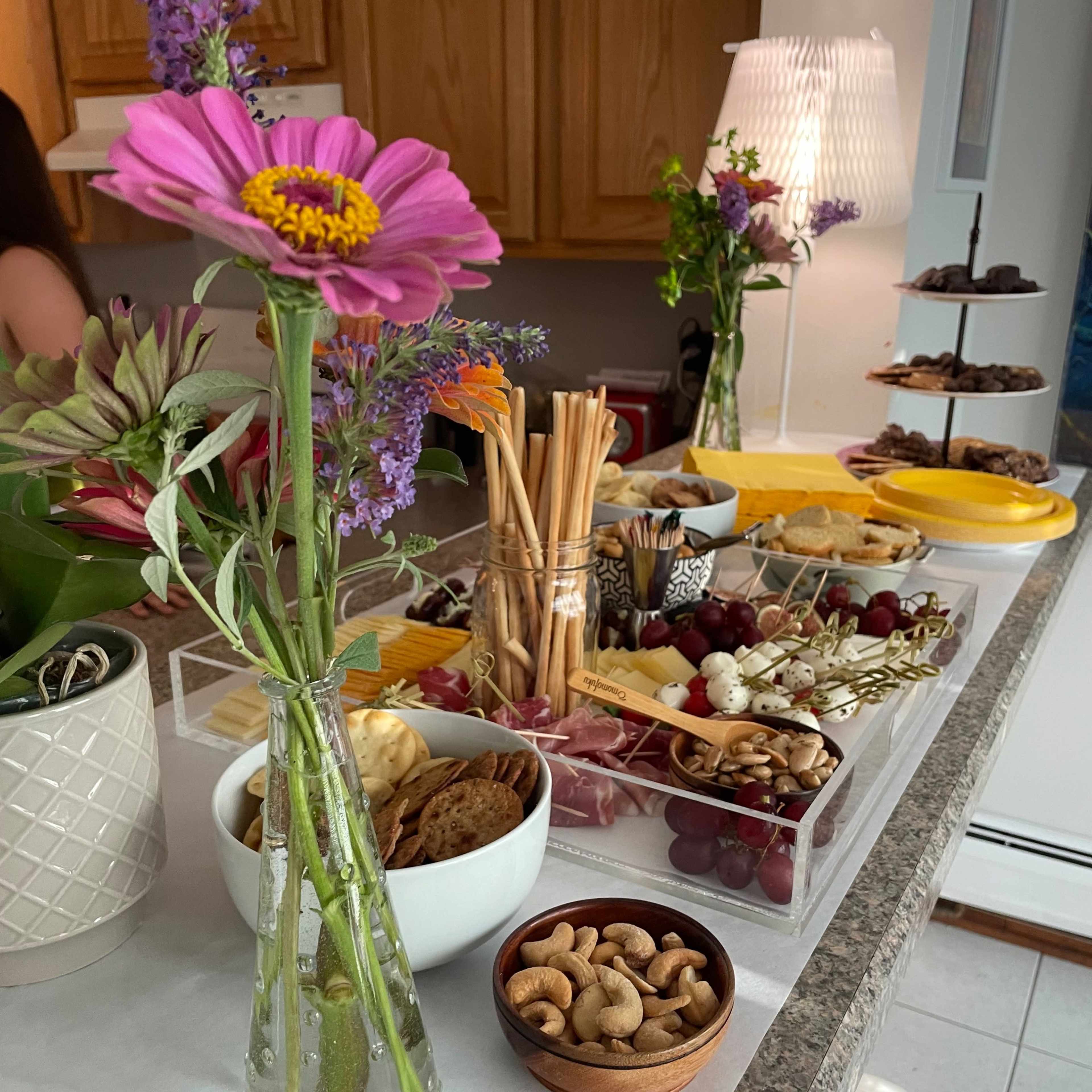 A variety of food items, including cheese, fruits, nuts, and snacks, is arranged on a kitchen counter alongside a decorative flower in a vase.