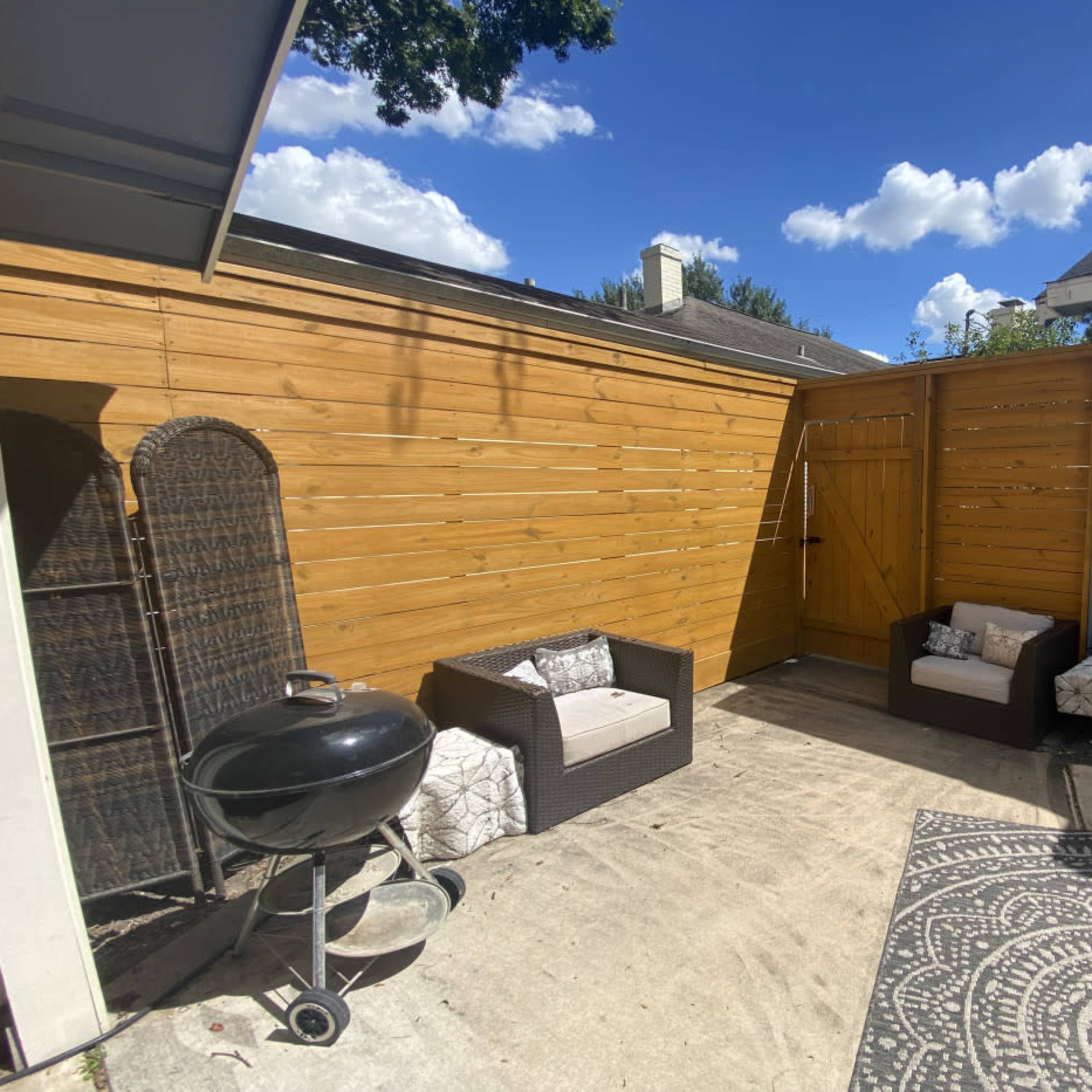 A small outdoor patio area featuring a wooden fence, two seating arrangements, a charcoal grill, and a decorative rug.