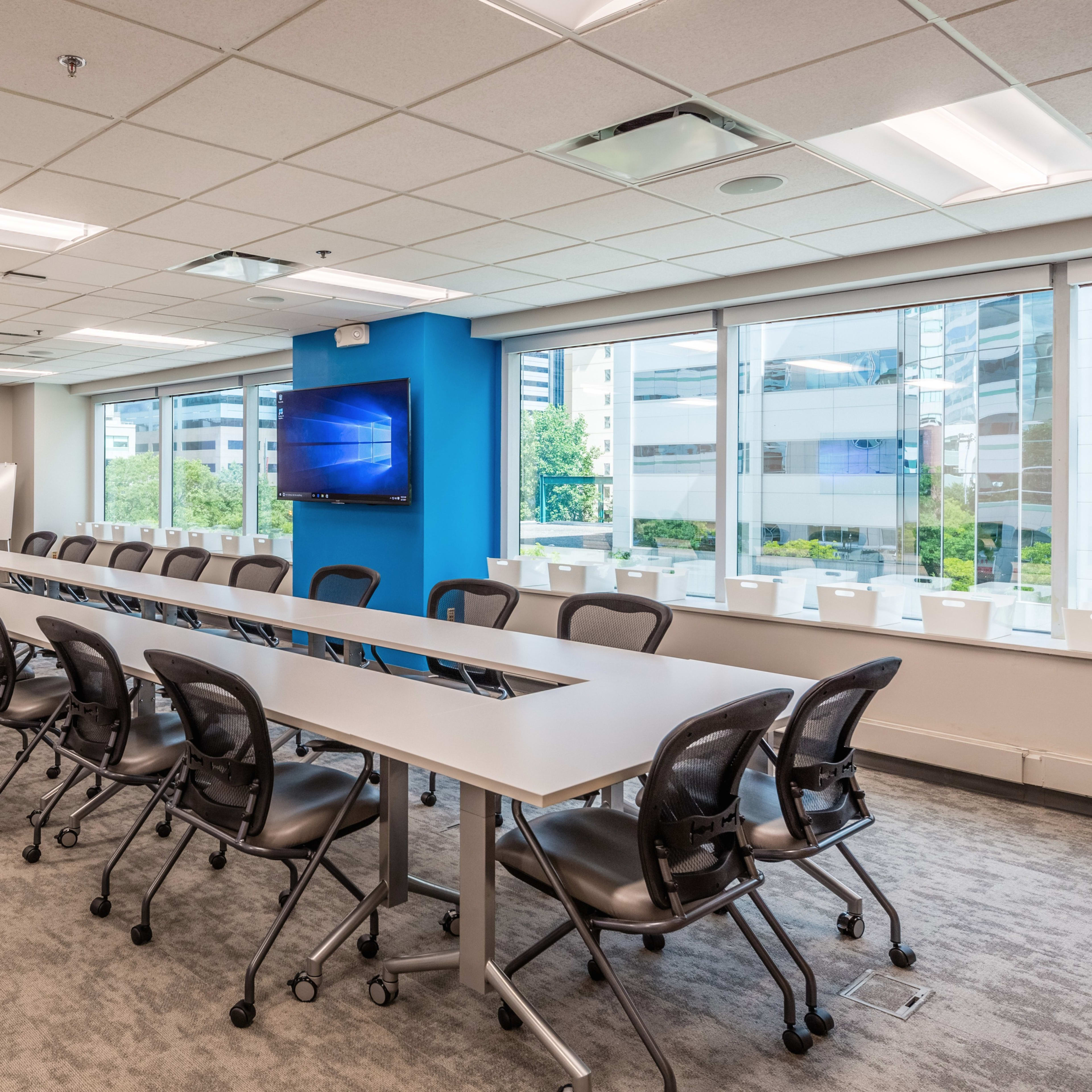 A modern conference room features a long table with rolling chairs, two large screens mounted on the walls, and windows that provide natural light.