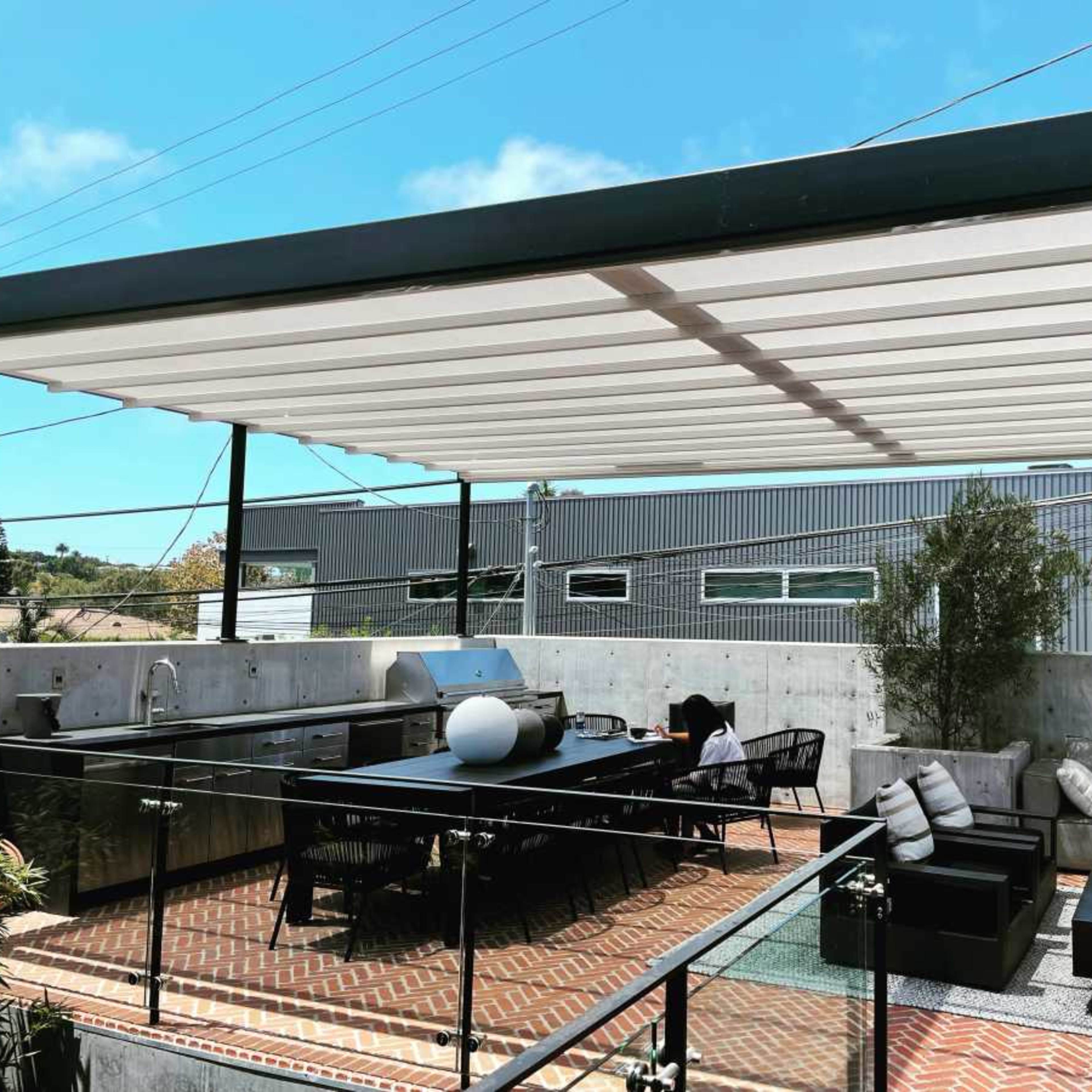 A rooftop terrace features a dining area with a table, sectional sofas, and an outdoor kitchen, surrounded by greenery and buildings in the background.
