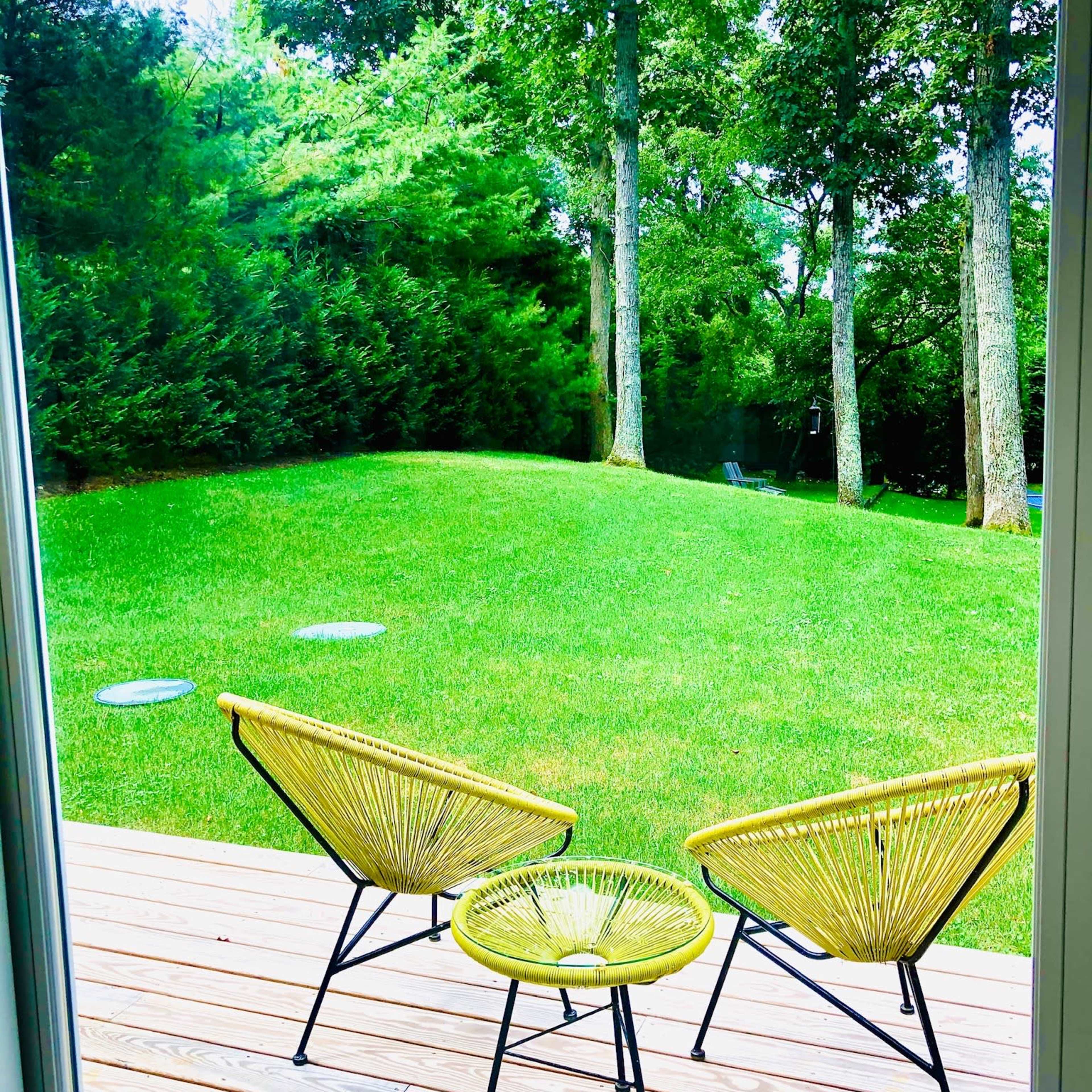 Two yellow chairs and a small table are positioned on a wooden deck overlooking a green lawn and trees.