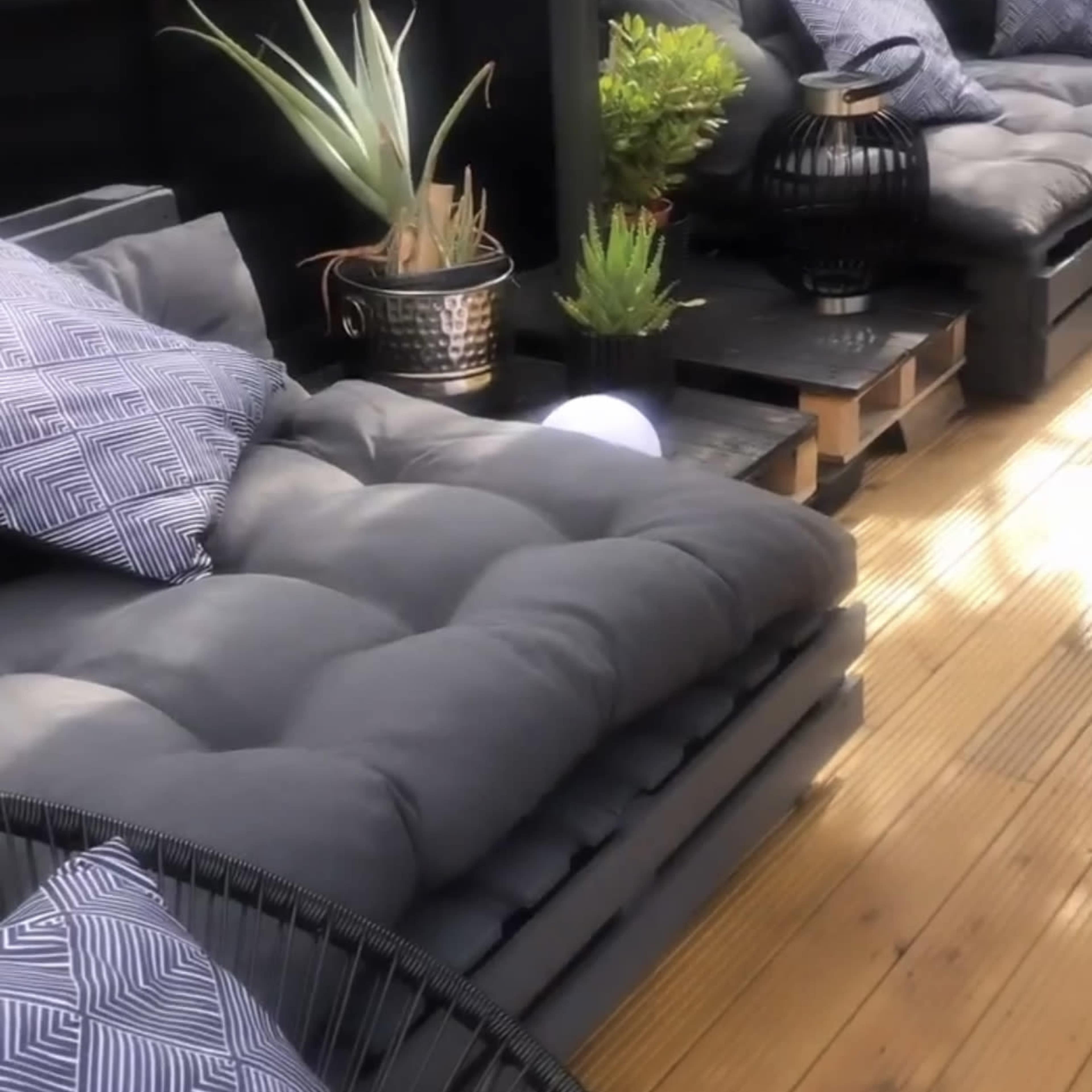 A cozy outdoor seating area featuring dark cushioned sofas, decorative plants, and accent pillows arranged on a wooden deck.